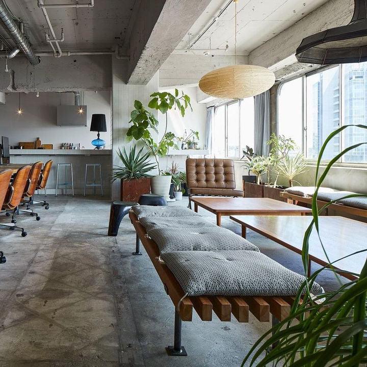 Wide loft seating view with a flattened paper shade by tall windows, plants and grey cushioned benches