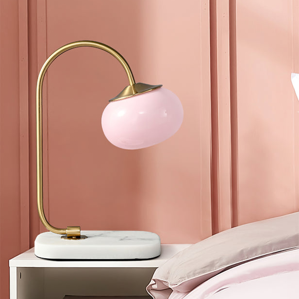 Pink globe shade on a gold-tone stem and pale veined base beside pink bedding against a panelled wall