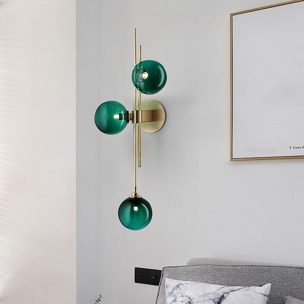 Green three-globe wall light on gold-tone rods above a grey headboard and patterned pillow near a gold-framed picture