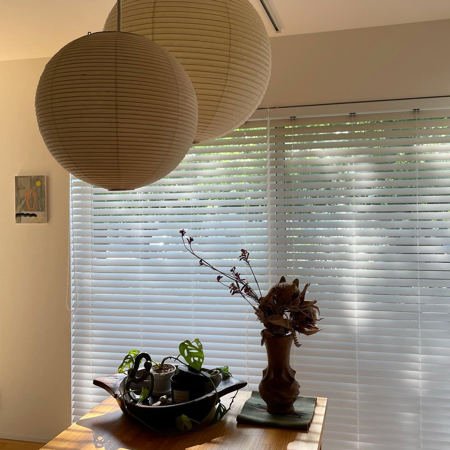 Two paper globes above a wood table with plants, wooden bowl and dried flowers before wide blinds