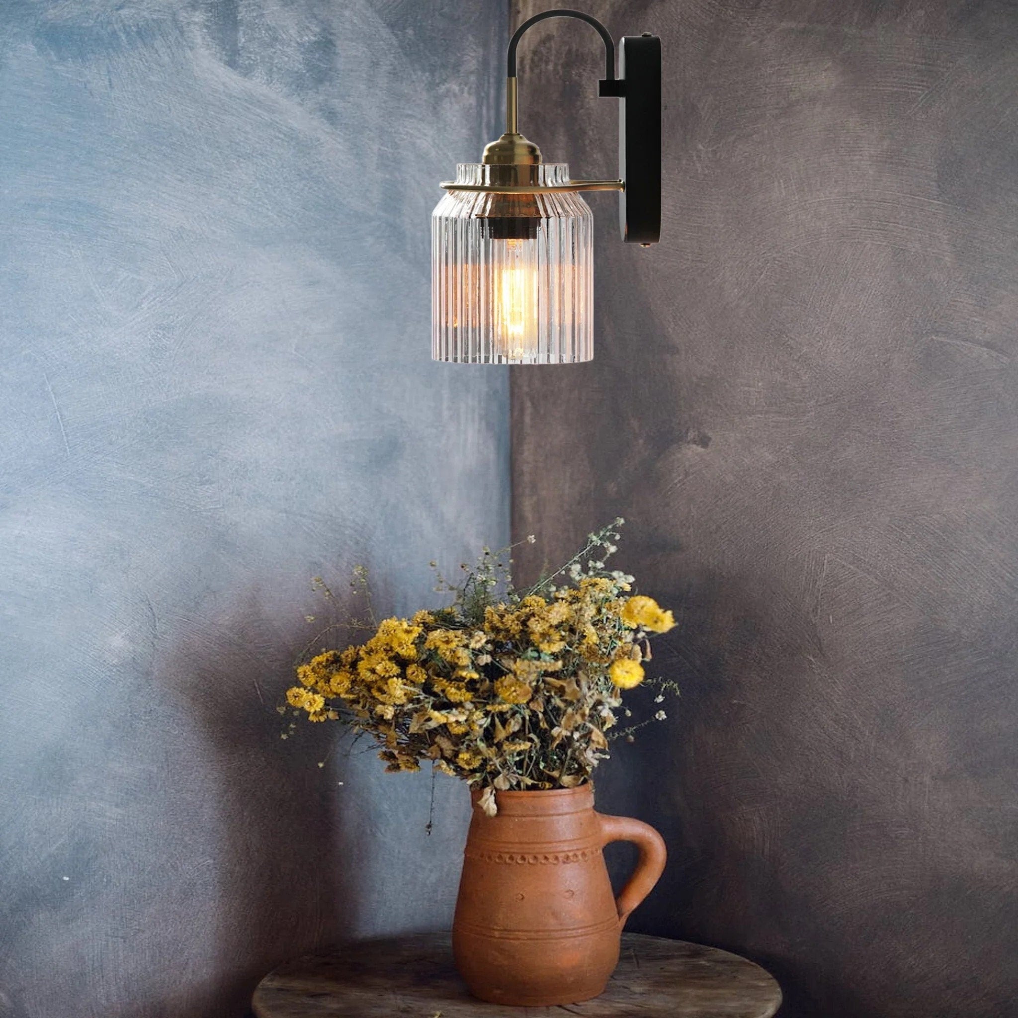 Downward glass wall light above a brown handled pitcher with yellow flowers on a round wooden table