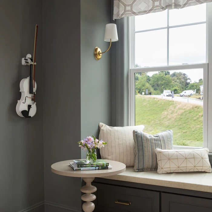 Single brass linen-shade wall light beside a window seat and round table.
