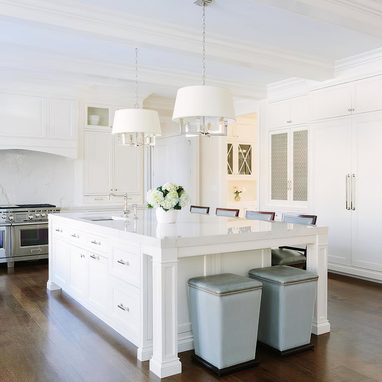 Two polished silver-looking shaded chandeliers above a white kitchen island with flowers and bar stools