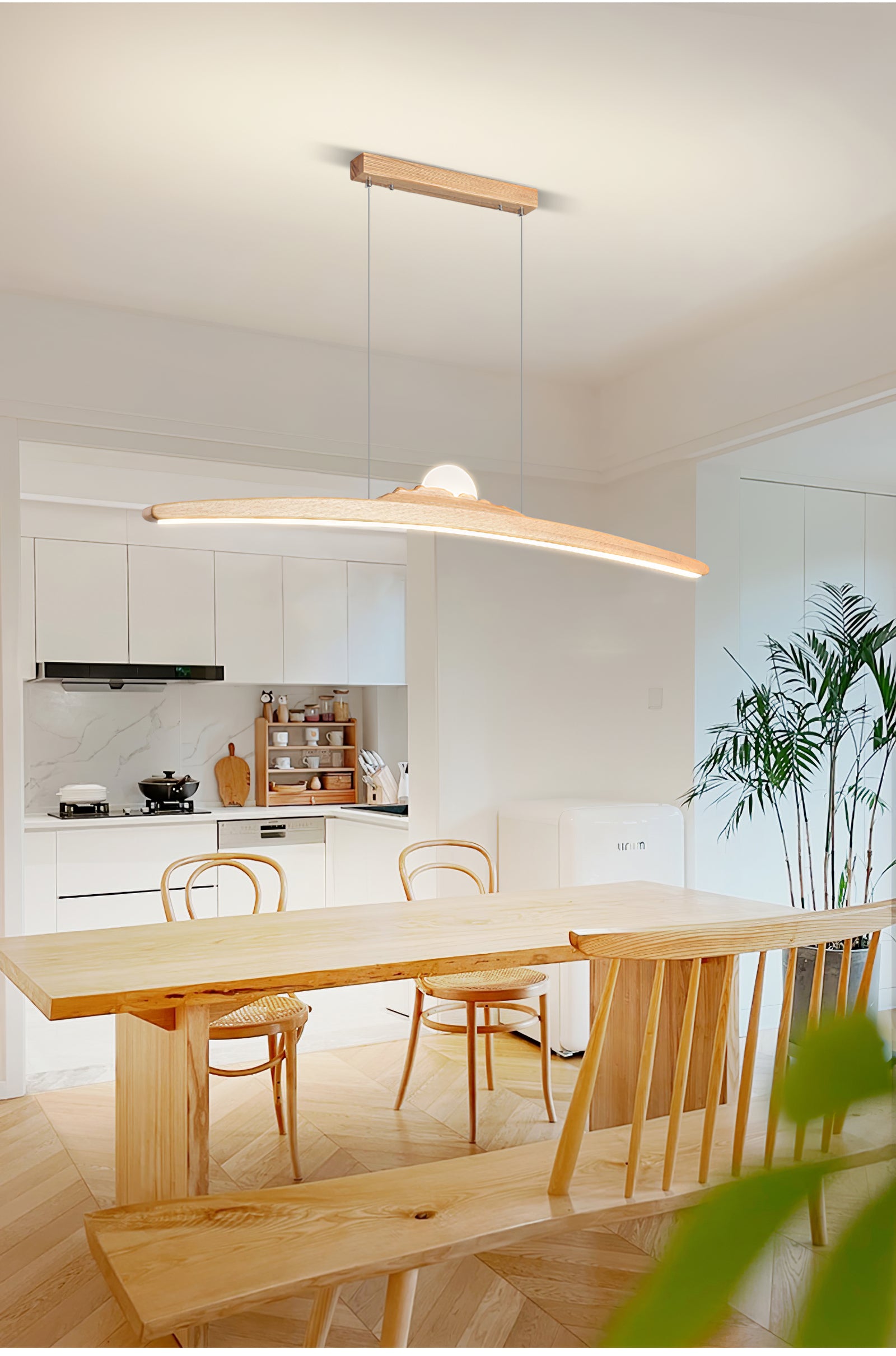 Pale wood-tone pendant over a timber table and woven chairs beside a white kitchen and potted palm