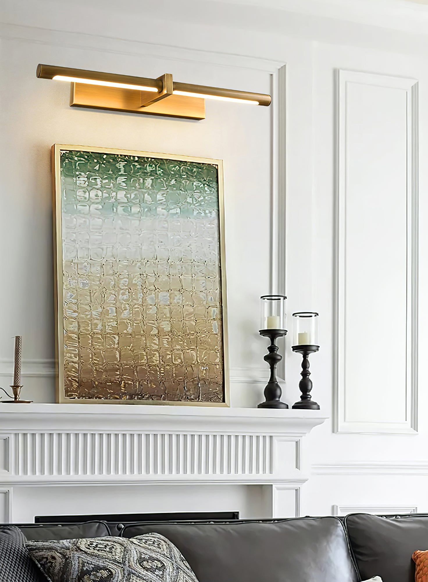 Lit brass-colour picture light above textured framed artwork on a mantel with candleholders