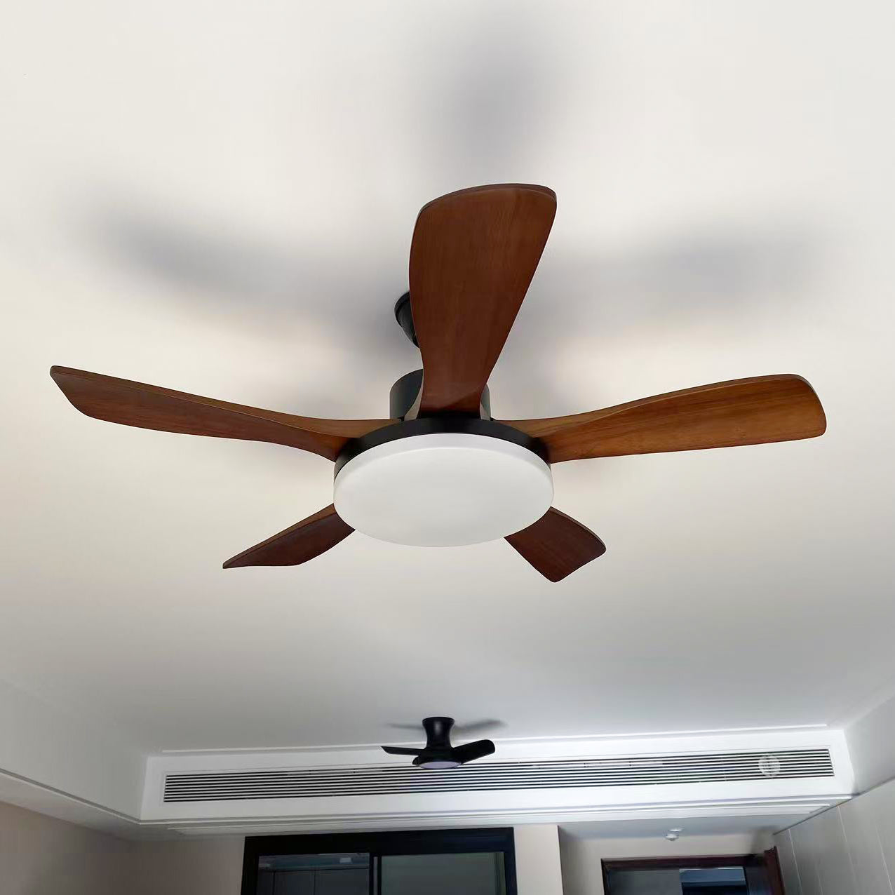 Dark brown five-blade ceiling fan with black housing and pale round light, viewed below with an air-conditioning grille behind