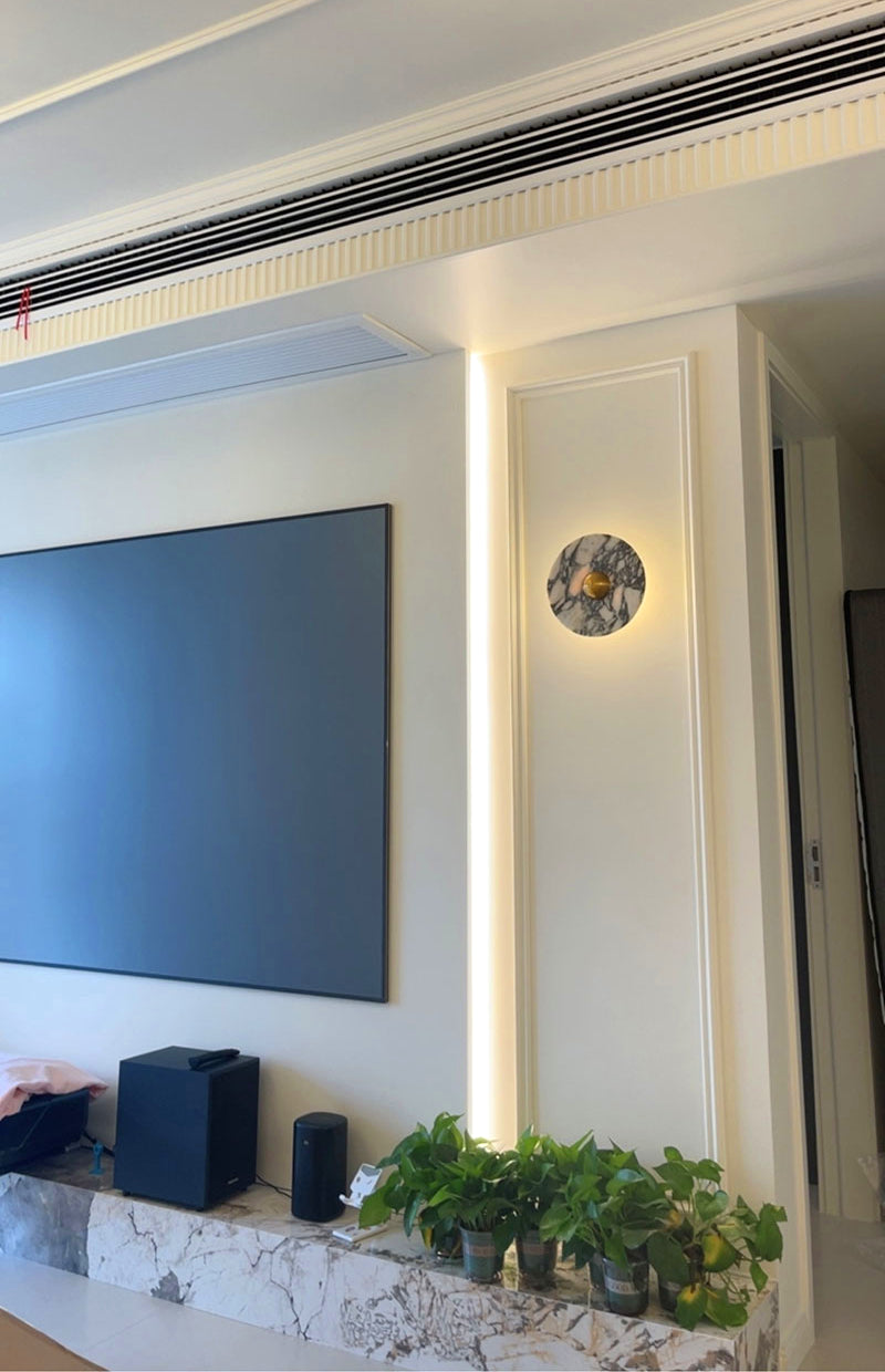 Gold-centred round stone wall light beside a TV, a narrow wall panel and plants on a shelf