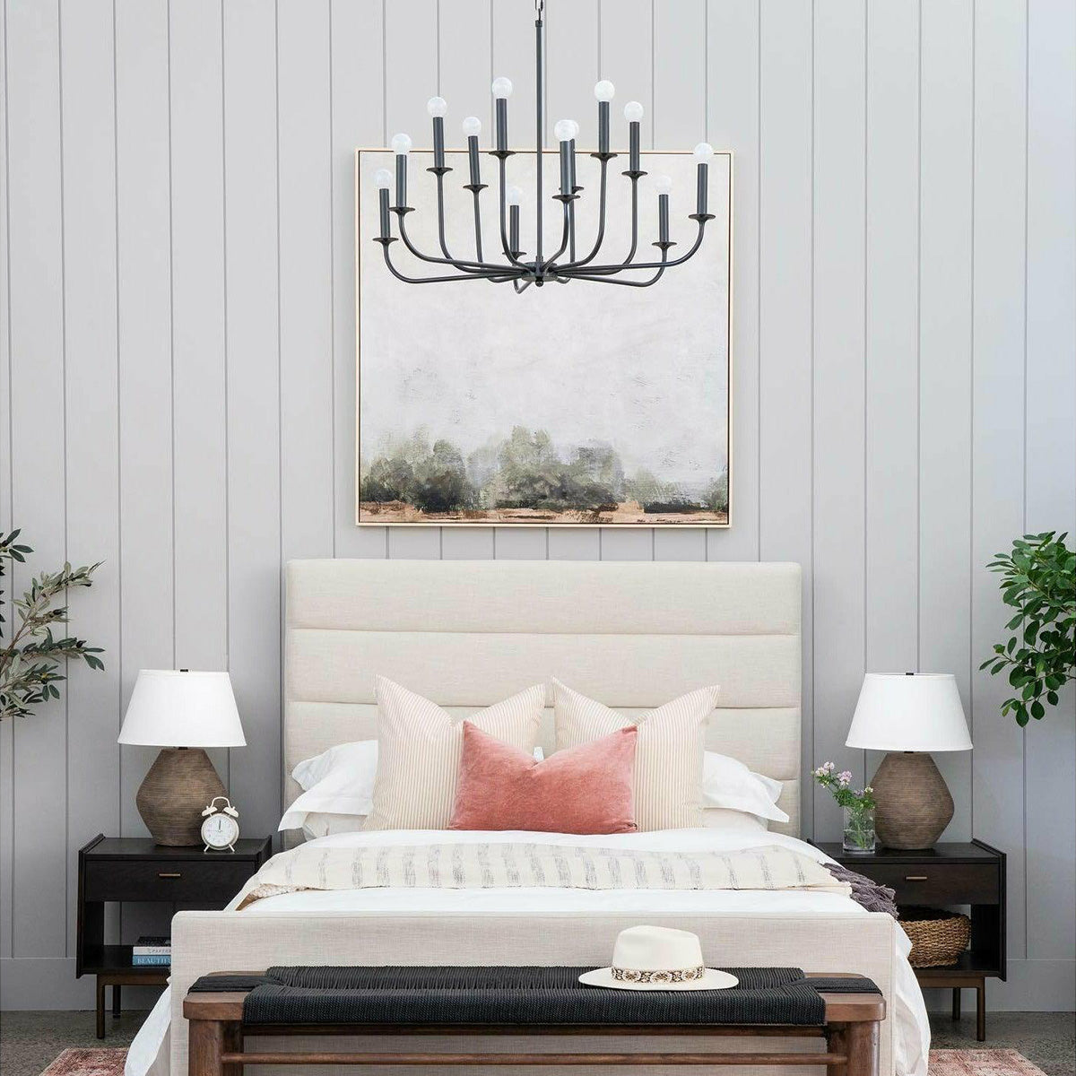 Black chandelier above a pale upholstered bed, with landscape artwork and separate lamps on bedside tables