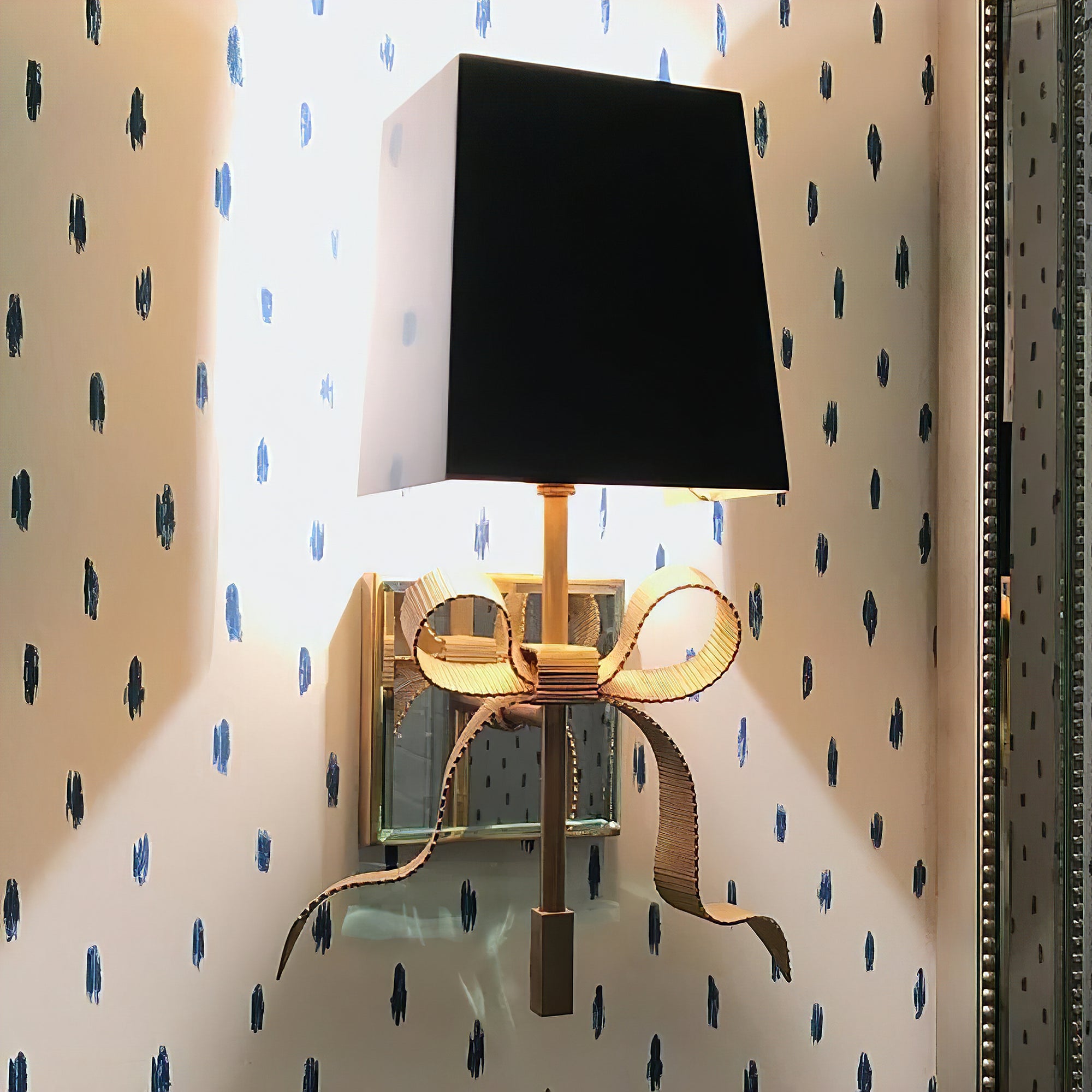 Close view of a black tapered shade above a textured brass-tone bow and reflective backplate on a dotted wall