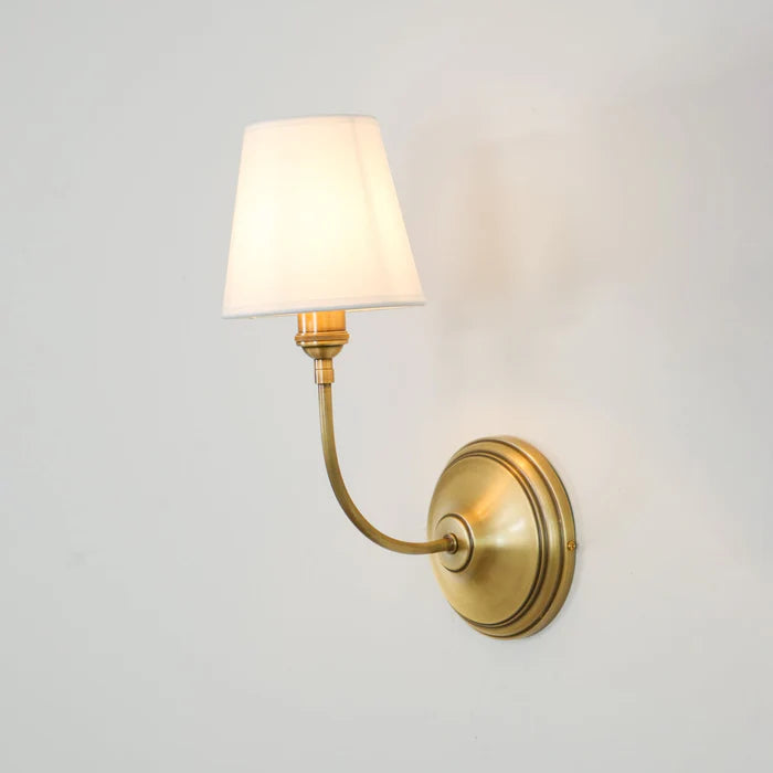 Angled view of a curved brass arm, round backplate and white linen shade.