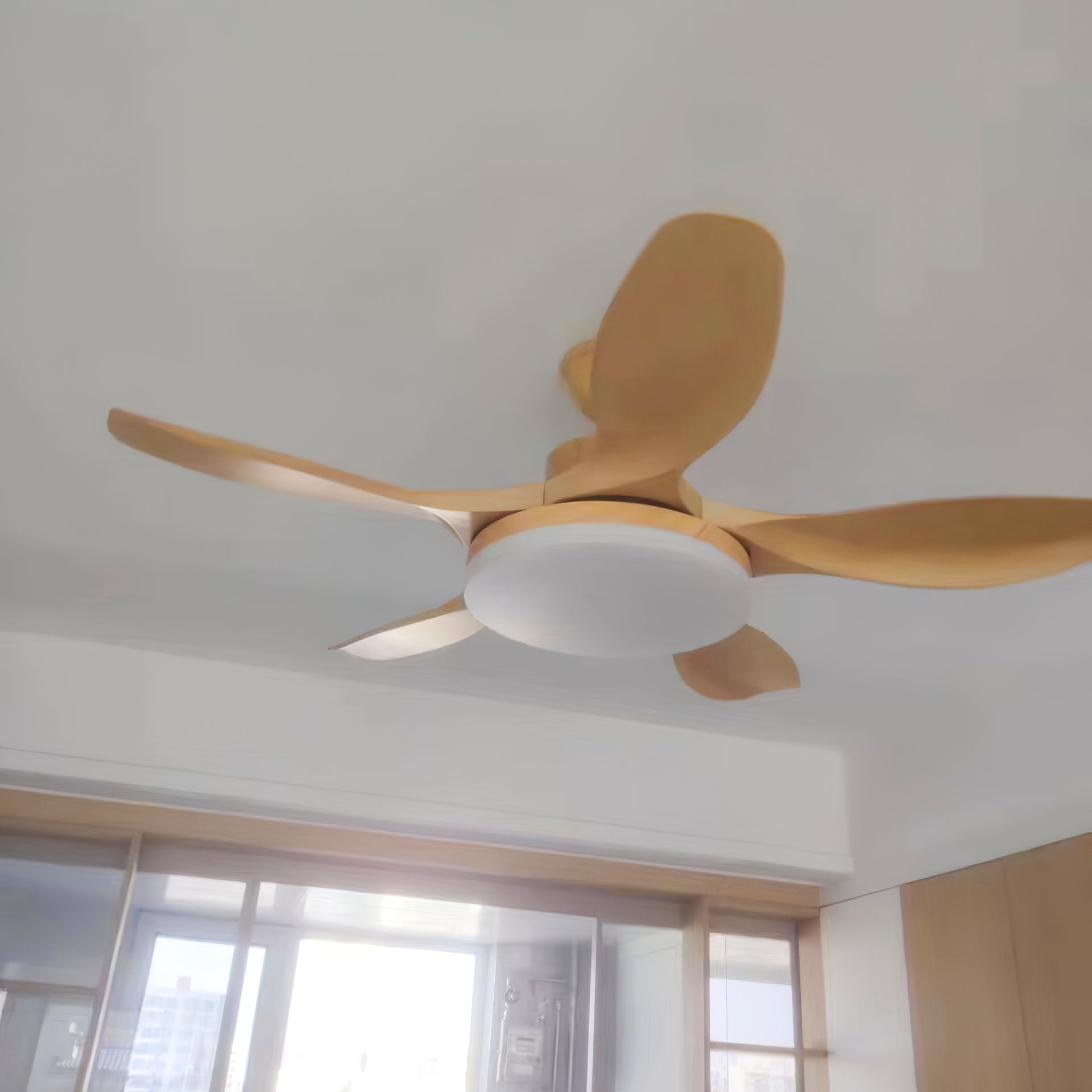 Close underside view of wood-tone curved fan blades and a round pale light near a bright window and wood cabinets