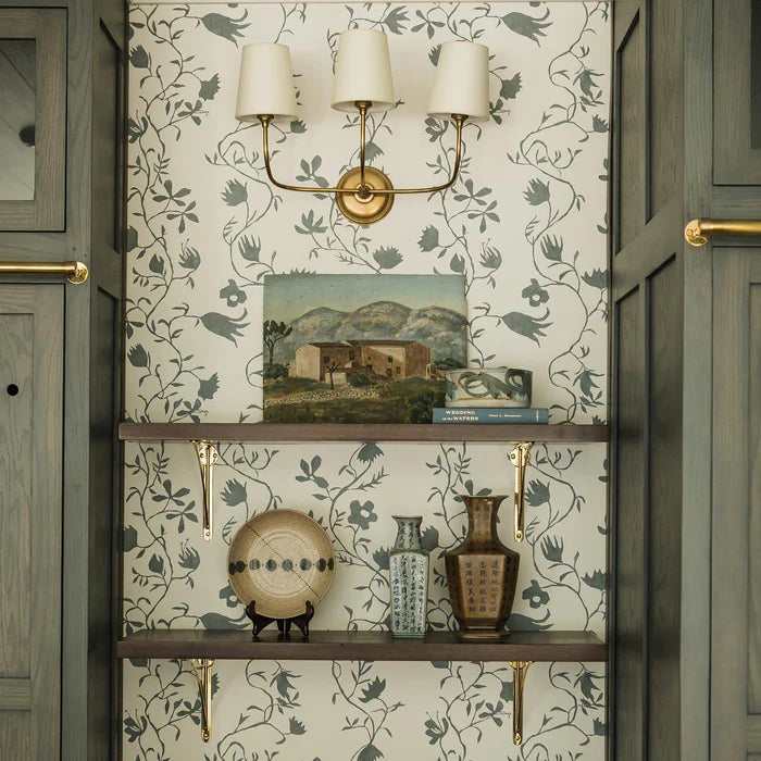 Brass two-shade wall light above shelves on a floral wall.