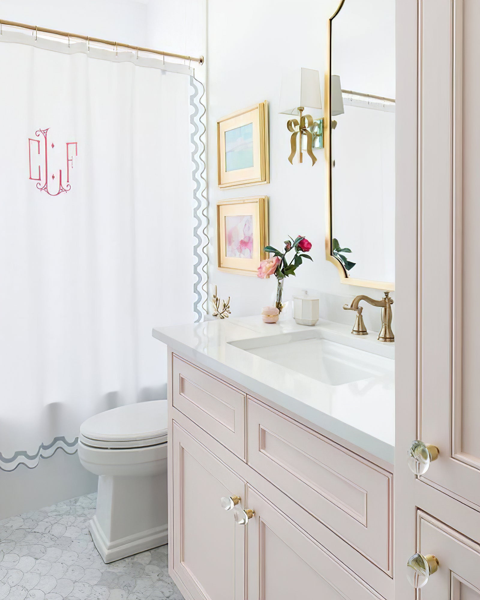Brass-tone bow light with pale shade beside a mirror above a pink cabinet, with framed pictures and a white curtain