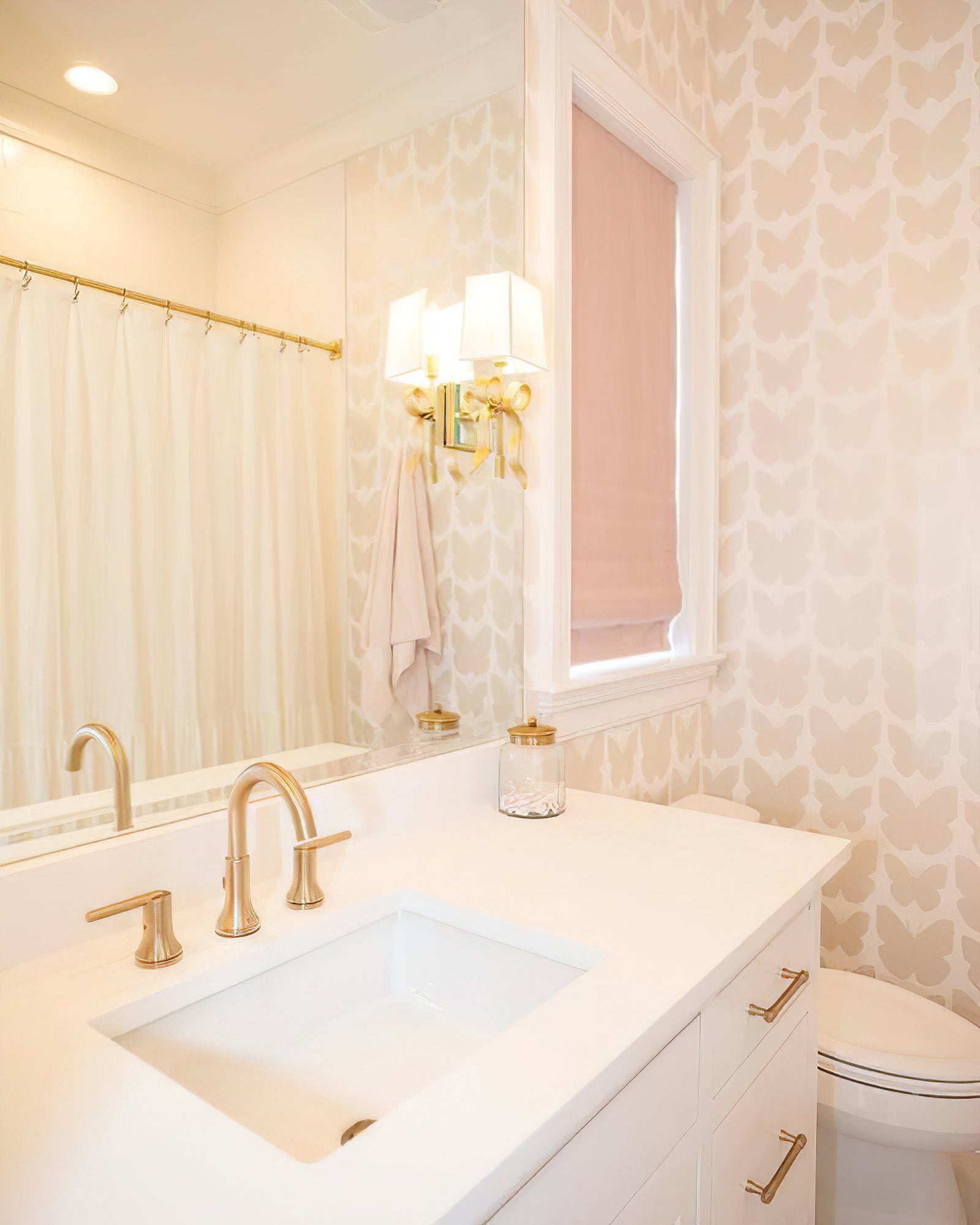 Lit brass-tone bow wall light and its reflection beside a pink window blind above a white sink