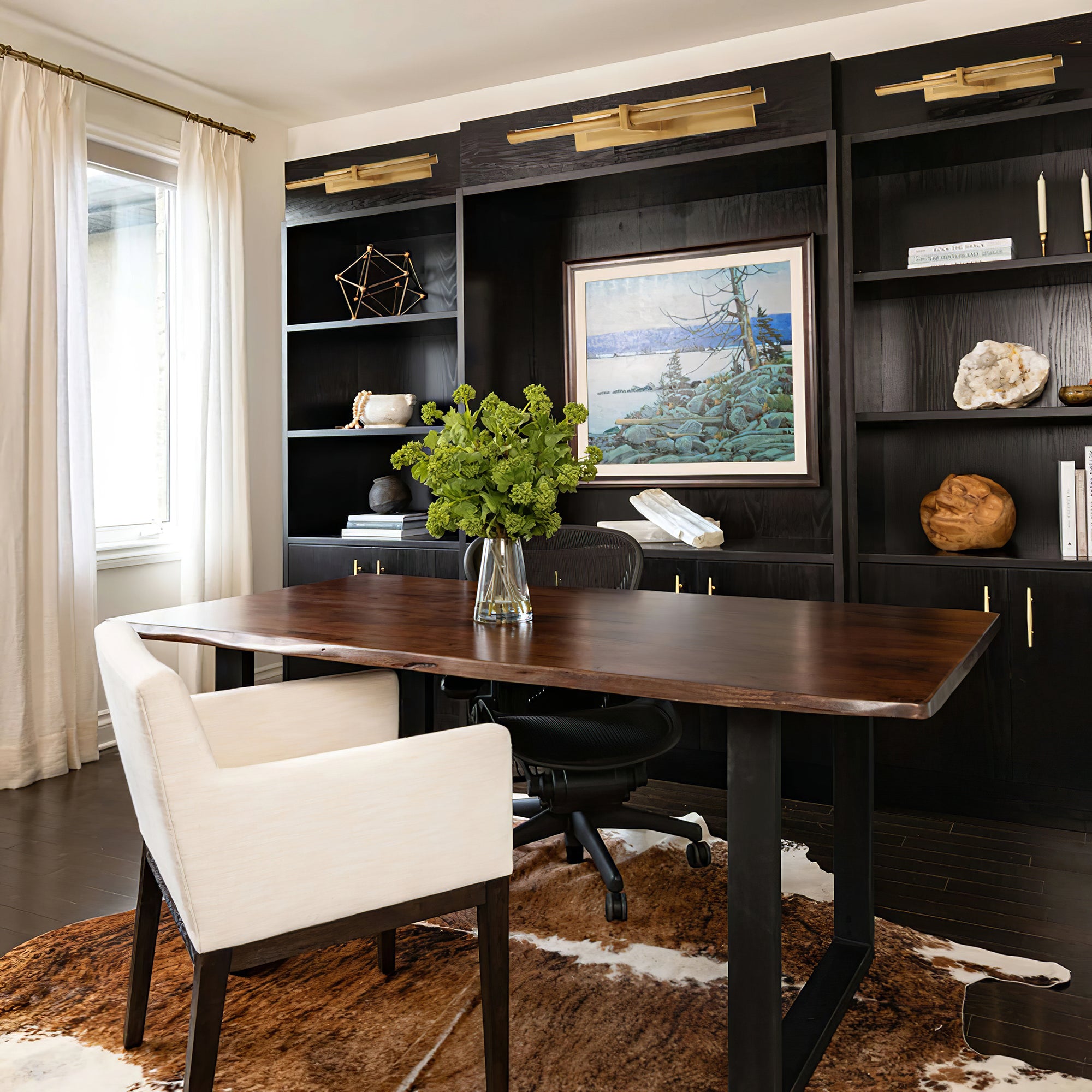 Brass-colour picture lights above black display shelving with framed art, sculpture and a wooden desk
