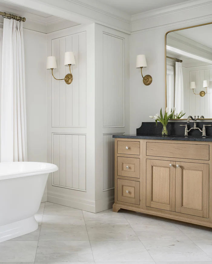 Single and double brass wall lights above a wood vanity near a bathtub.