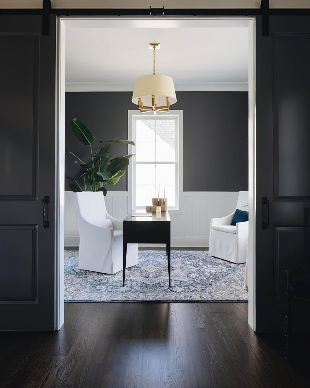 Gold-toned shaded chandelier above a dark desk and two white chairs, viewed through black sliding doors