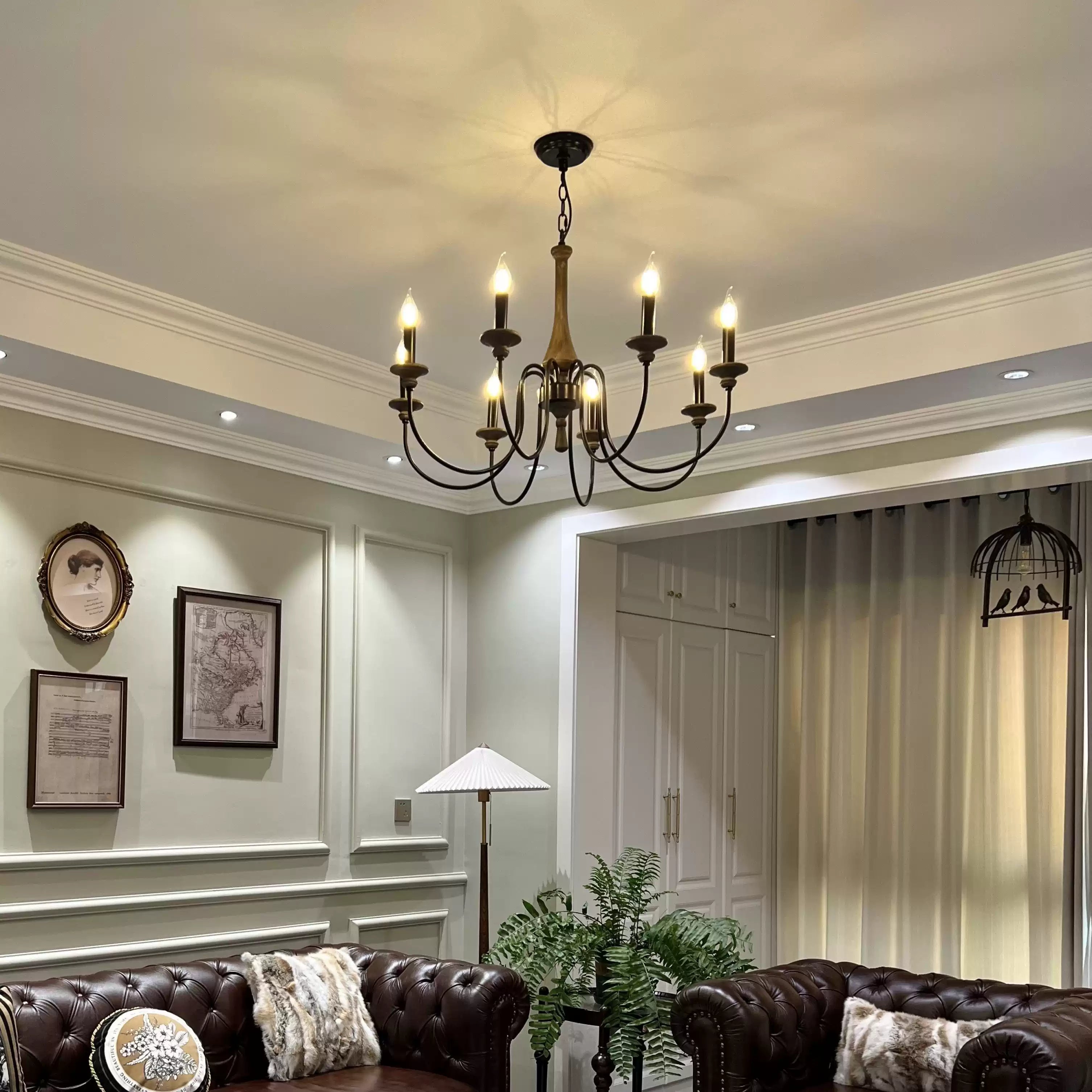 Wood-toned chandelier above brown tufted sofas beside framed art, a fern and separate floor lamp