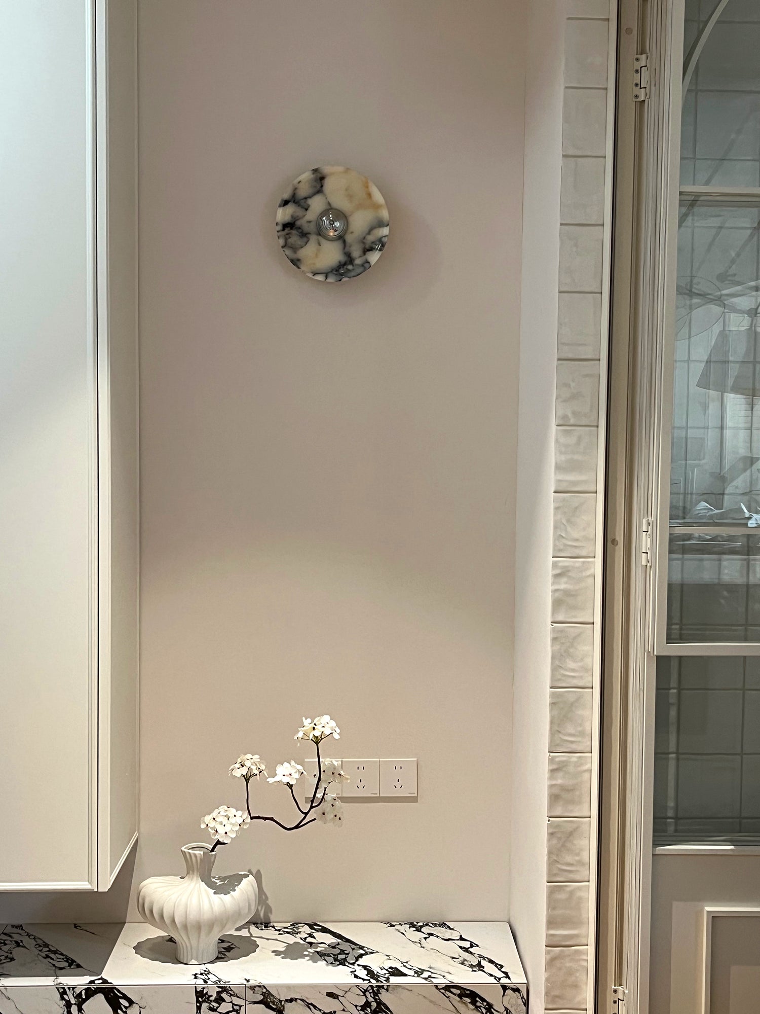 Silver-centred veined stone wall light above a patterned shelf with a white vase and flowering branches