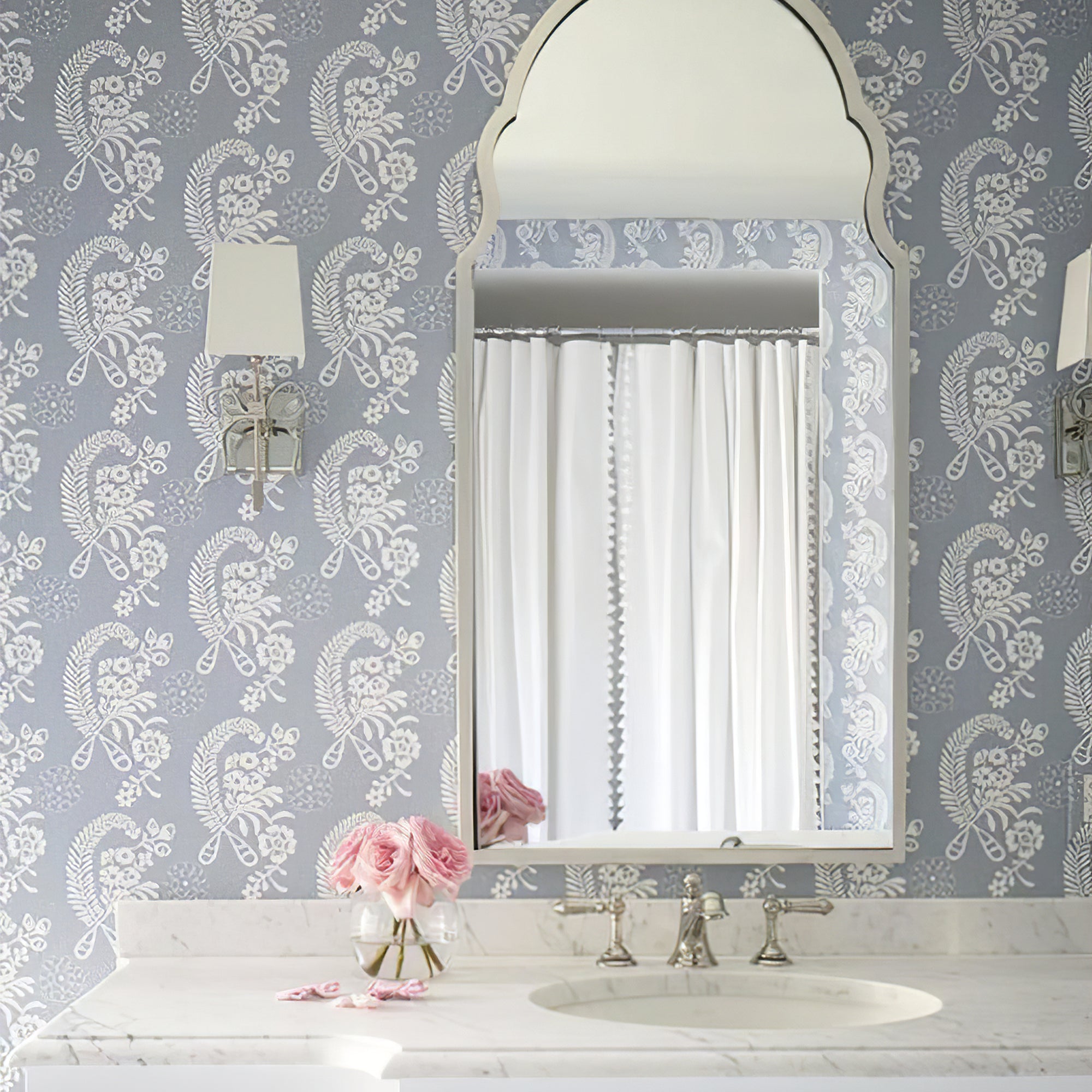 Silver-tone bow wall light beside a shaped mirror on blue floral wallpaper, with a second light partly visible at right