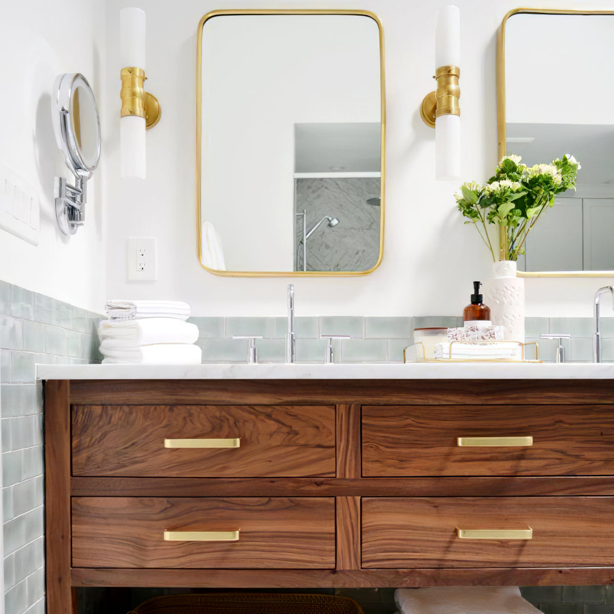 Brass-tone double cylinder wall lights beside gold-trimmed mirrors over a wood vanity with white flowers