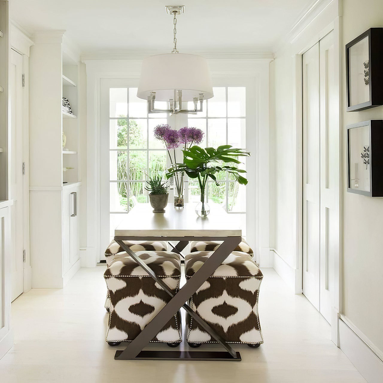 Polished silver-looking shaded chandelier above an X-legged table with upholstered stools and tall windows