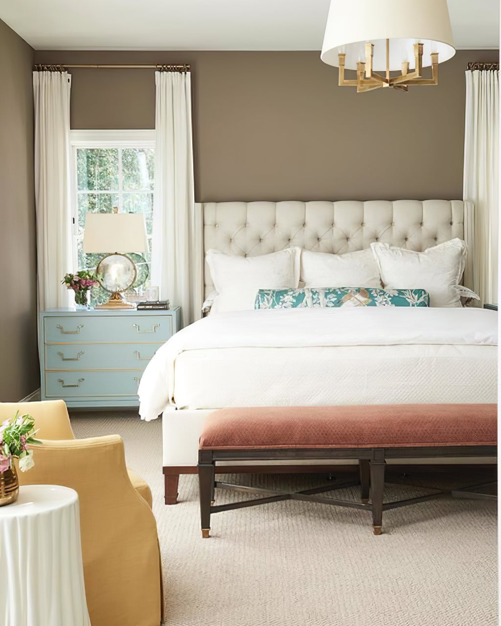 Gold-toned shaded chandelier partly visible above a cream tufted bed, teal bedside drawers and a red bench