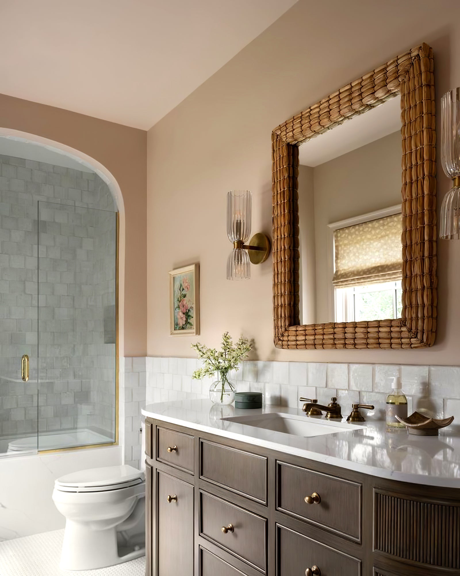 Pair of brass-tone double glass sconces beside a woven-look mirror over a brown vanity against a pink wall