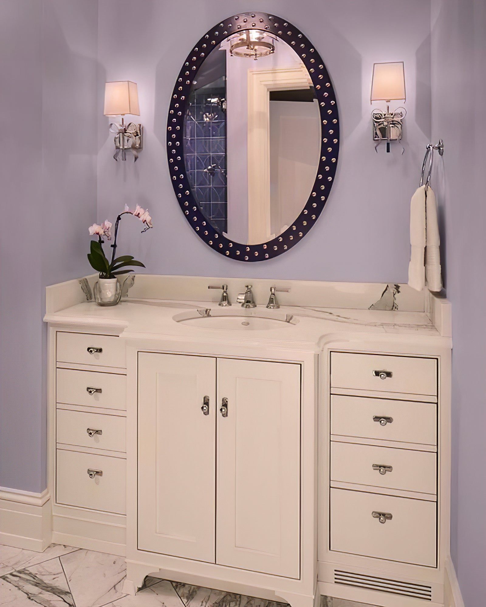 Two illuminated pale-shade silver-tone bow lights beside a dark oval mirror above a white sink cabinet