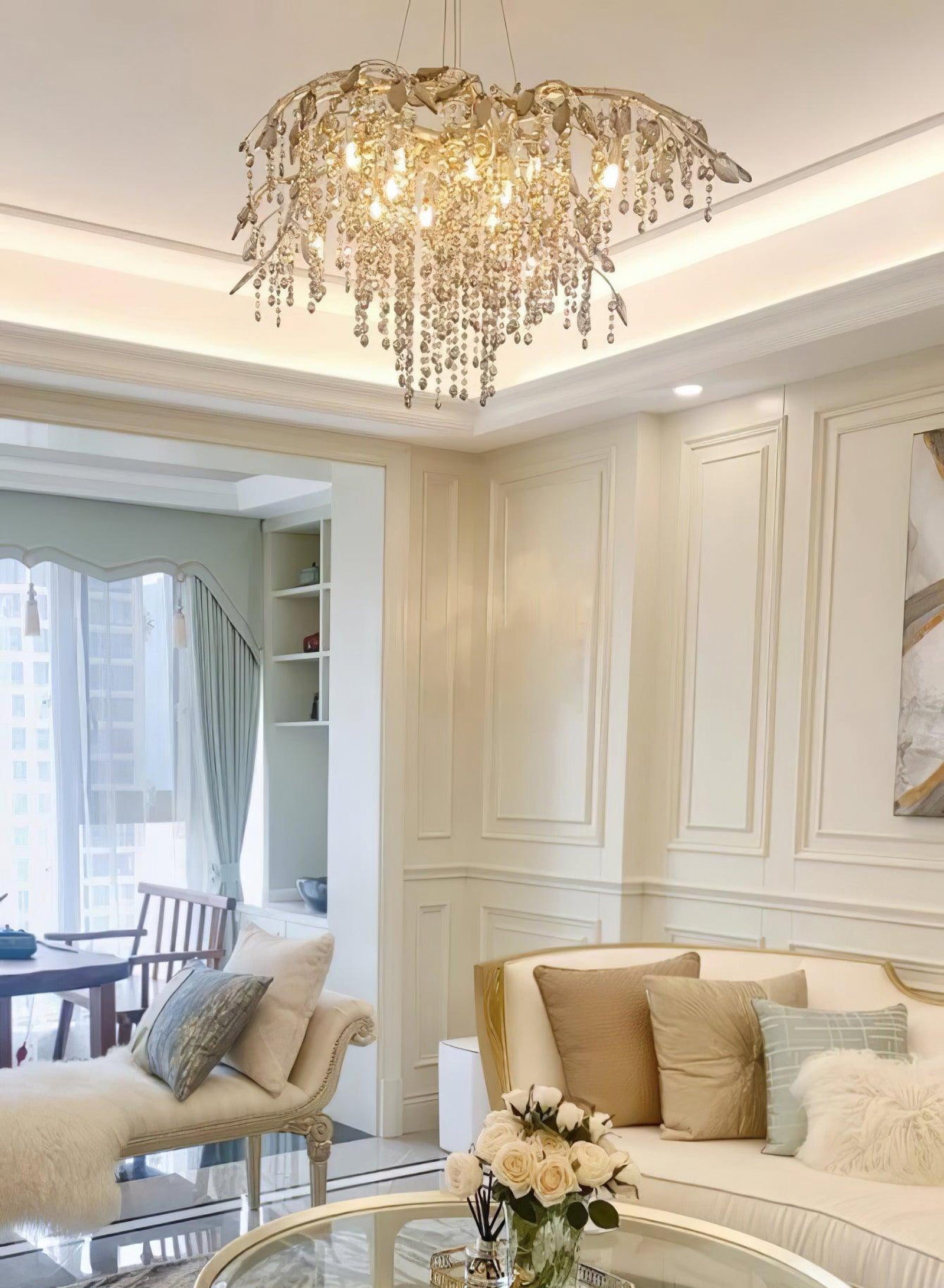 Glowing silver-tone branch chandelier above cream sofas and a glass table with flowers in a panelled room