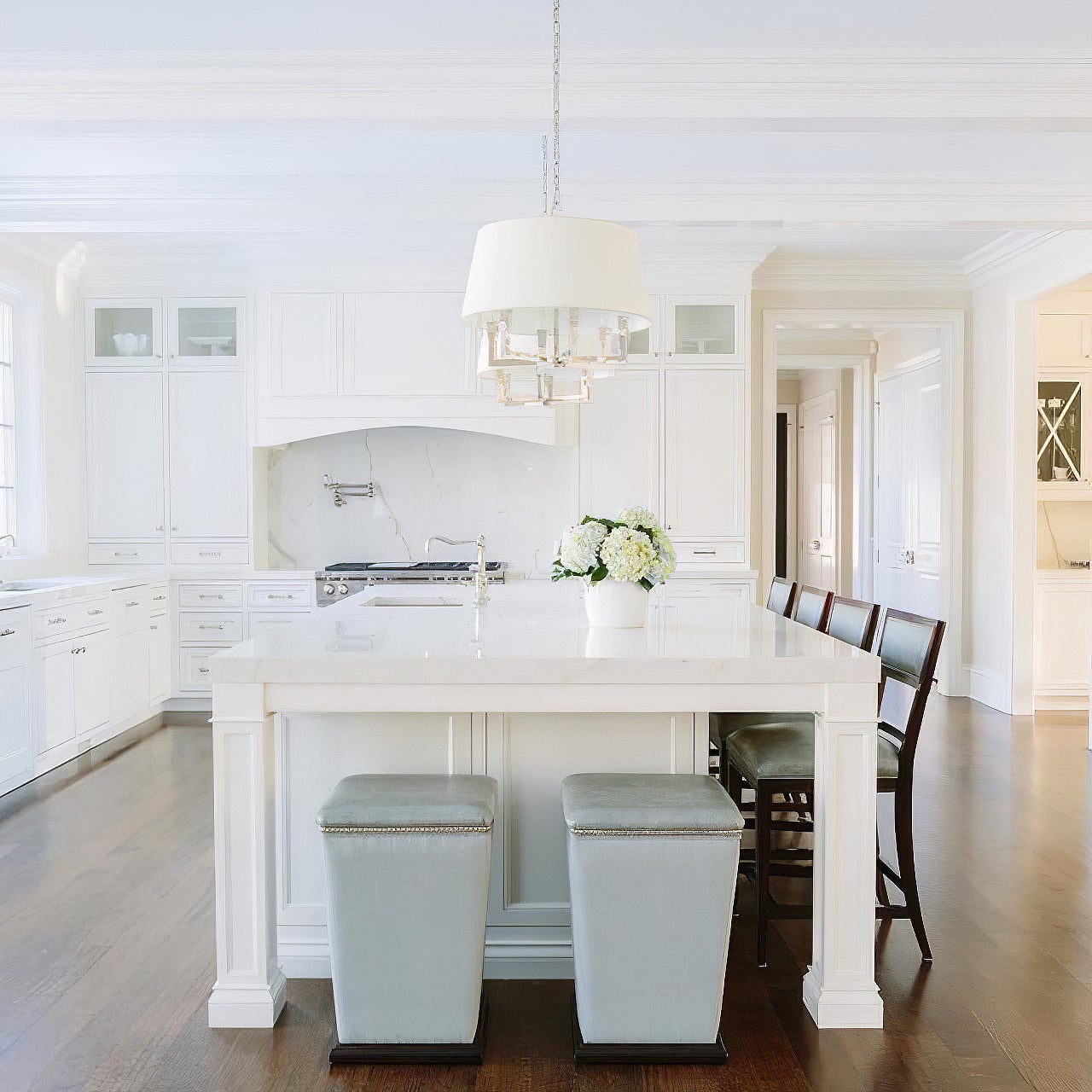 Silver-looking shaded chandeliers aligned over a white kitchen island, with flowers and stools to the right
