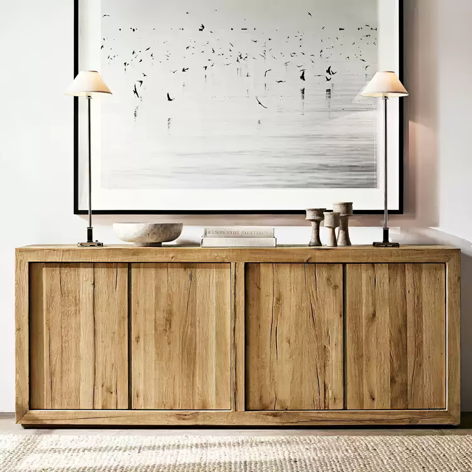 Two black-stem Slim Candlestick lamps with light shades flanking books and stone objects on wooden sideboard beneath artwork
