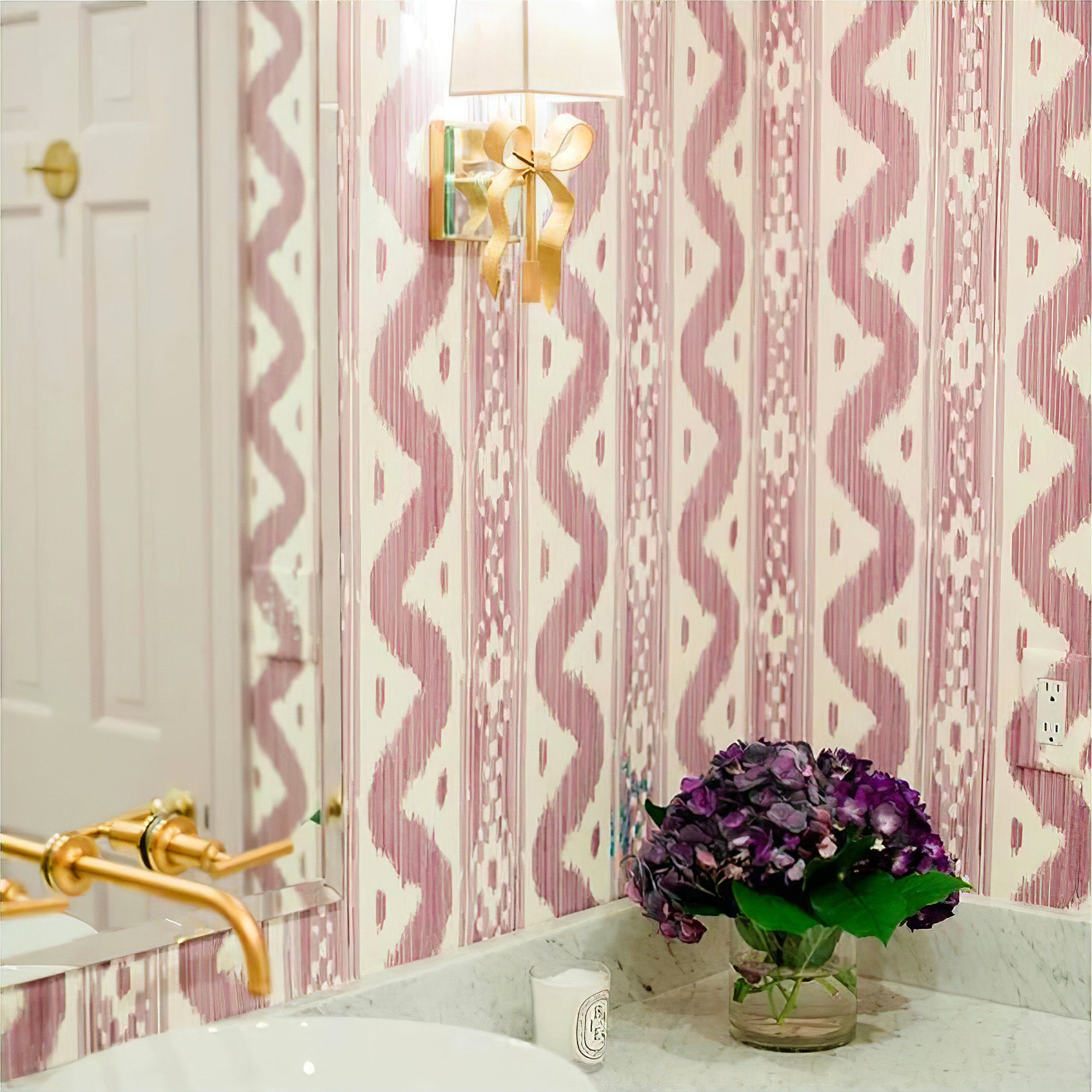 Brass-tone bow wall light with pale shade on pink patterned wallpaper beside a mirror and purple flowers