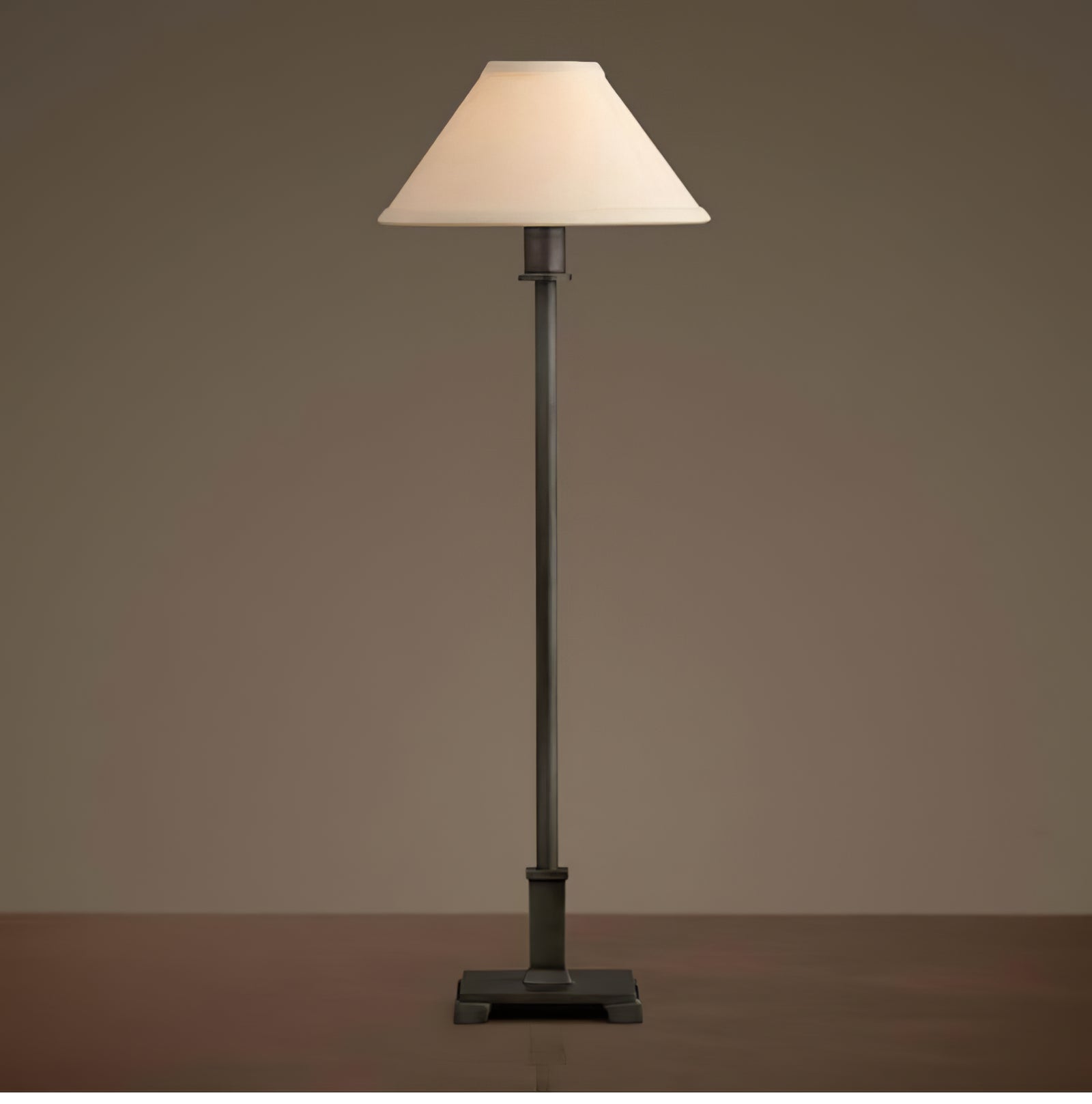 Dark-stem Slim Candlestick table lamp with glowing cream shade and square stepped base on dark wood against neutral background