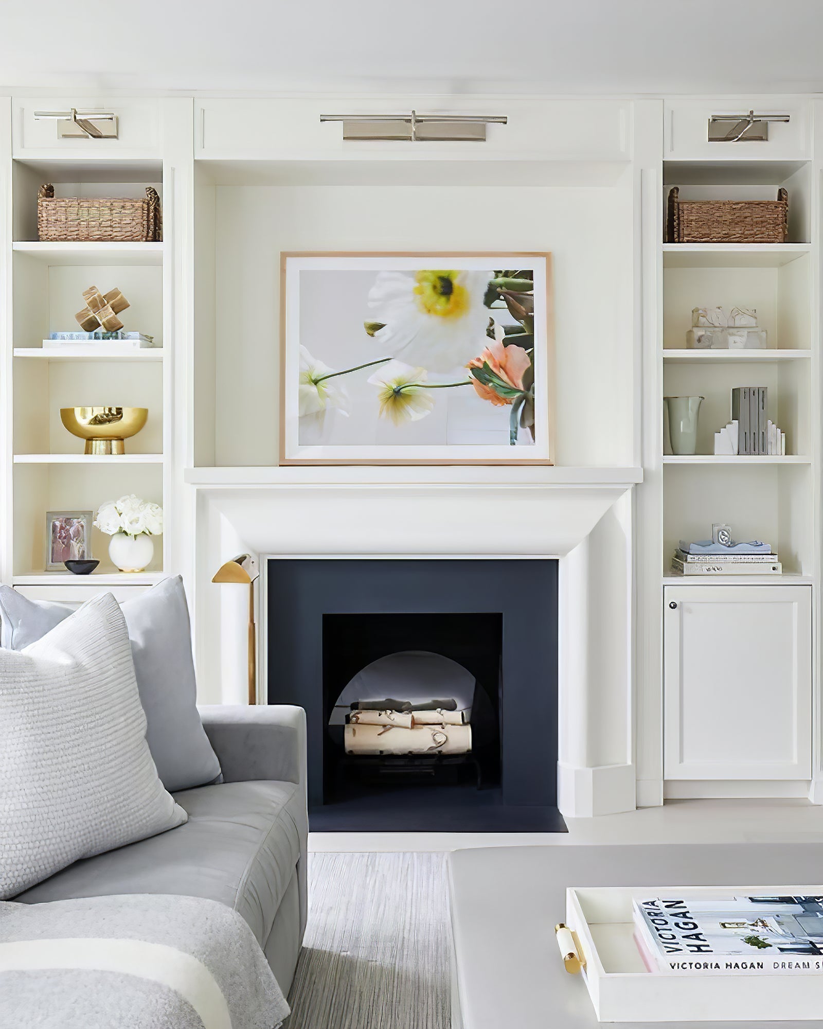 Silver-colour picture lights above white built-ins around a fireplace with floral art and a gray sofa
