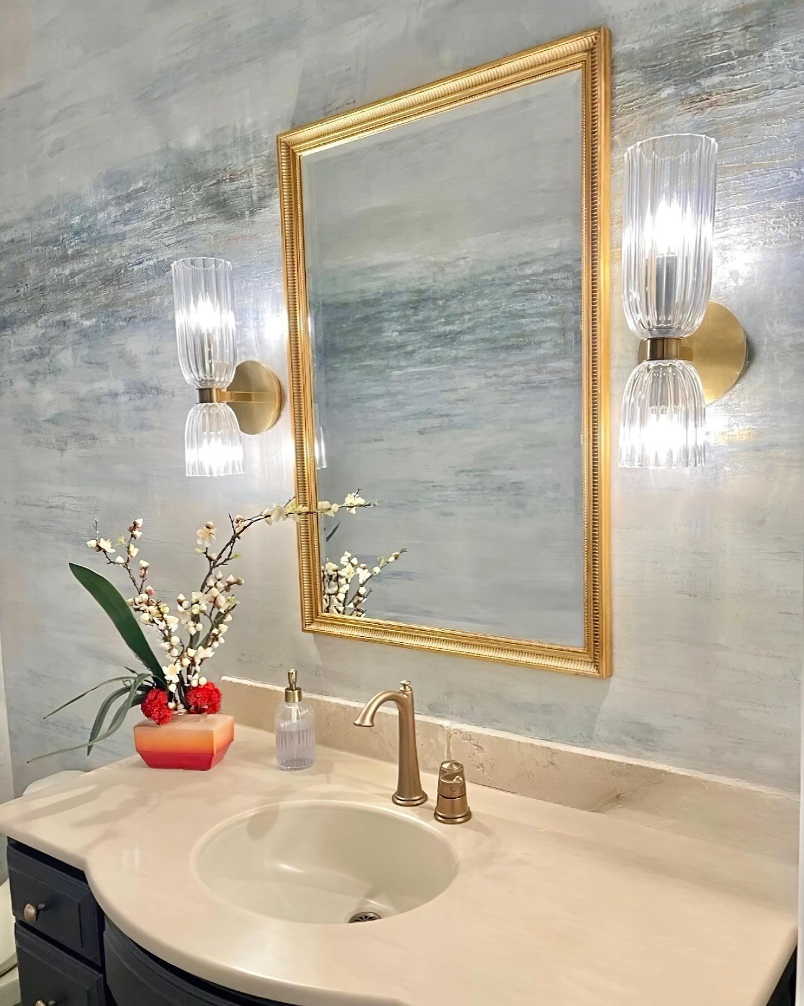 Two glowing brass-tone double glass sconces flanking a rectangular gold-framed mirror above a basin