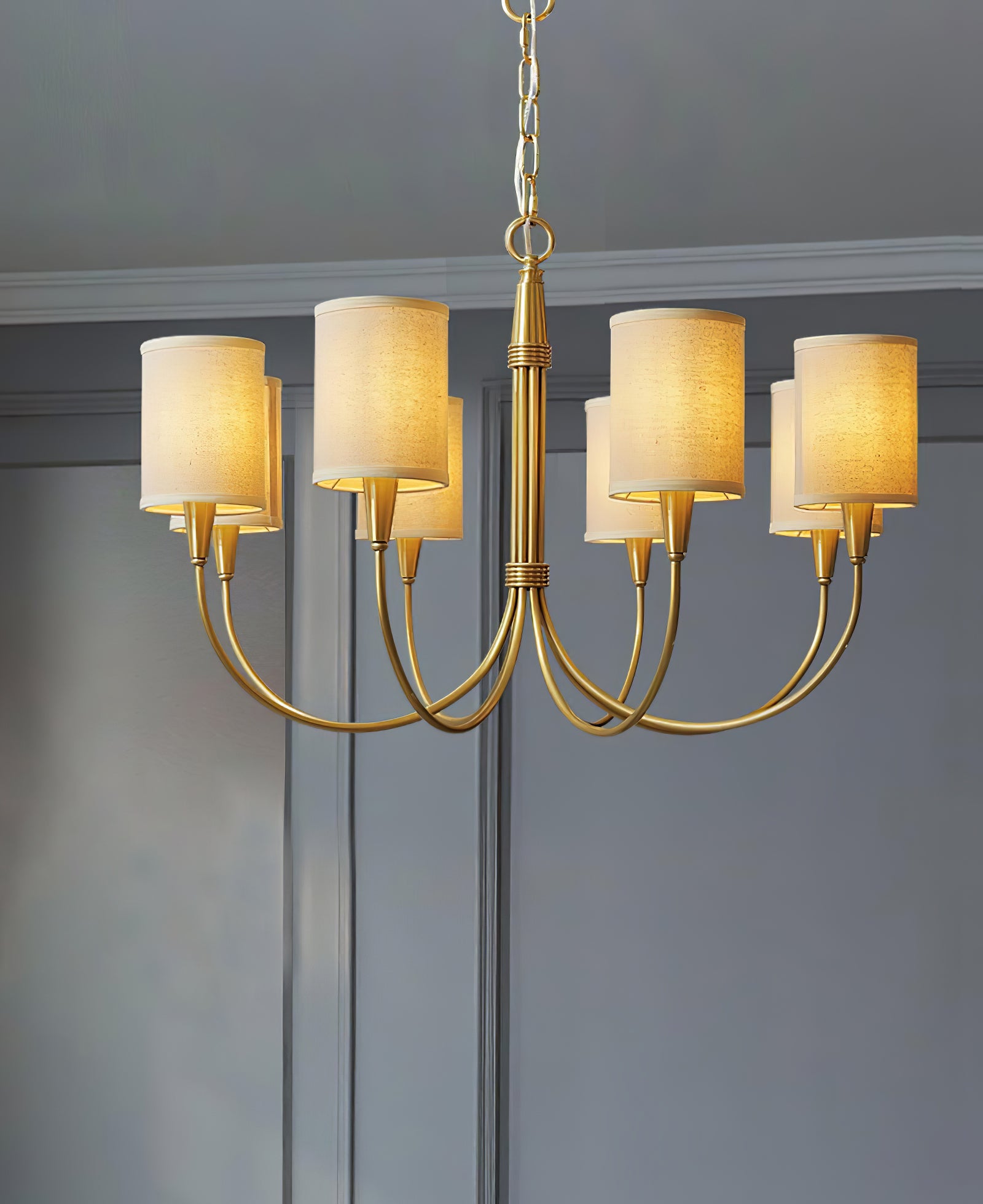 Eight-light Quincy chandelier with beige cylindrical shades and gold-tone curved arms before a grey panelled wall