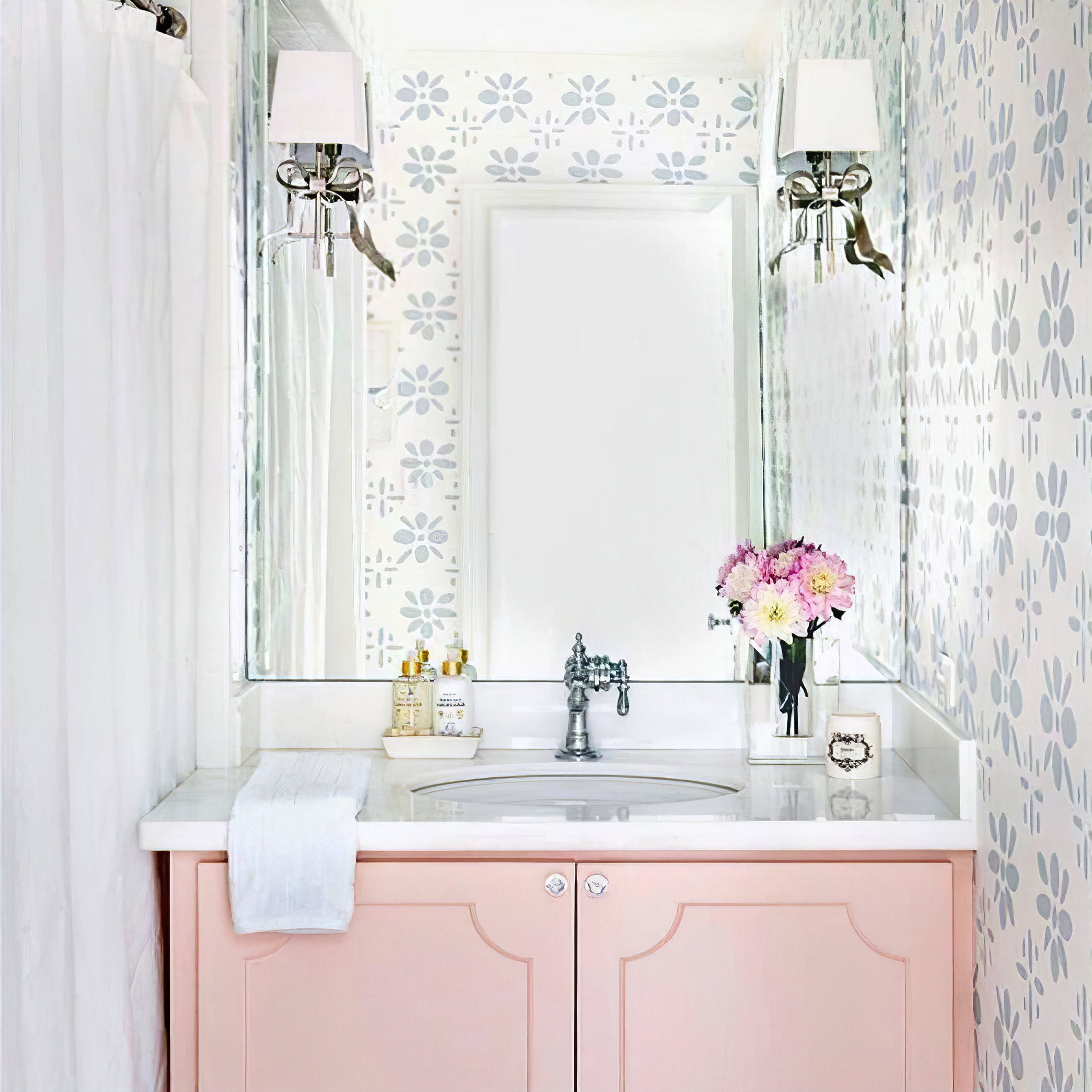 Silver-tone bow wall lights with pale shades beside a mirror above a pink sink cabinet and floral wallpaper