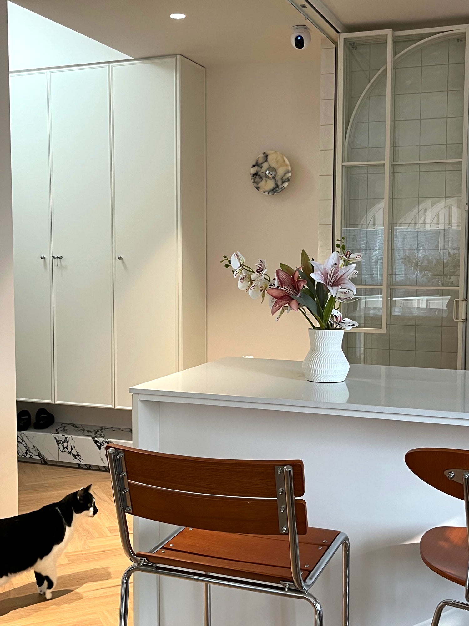 Silver-centred stone wall light behind lilies on a white counter, with dining chairs and a cat in the foreground