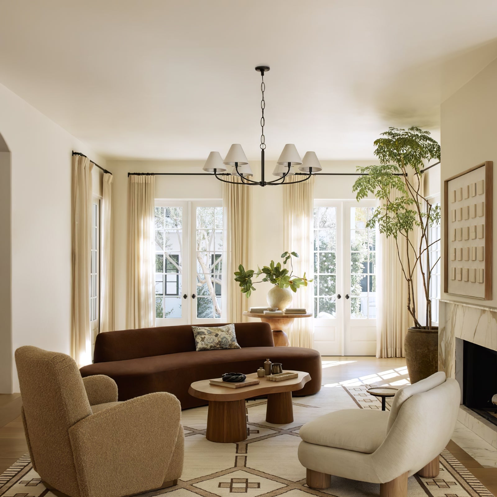 Black Stacey chandelier with white fabric shades above living room sofa and coffee table beside tall plant and fireplace