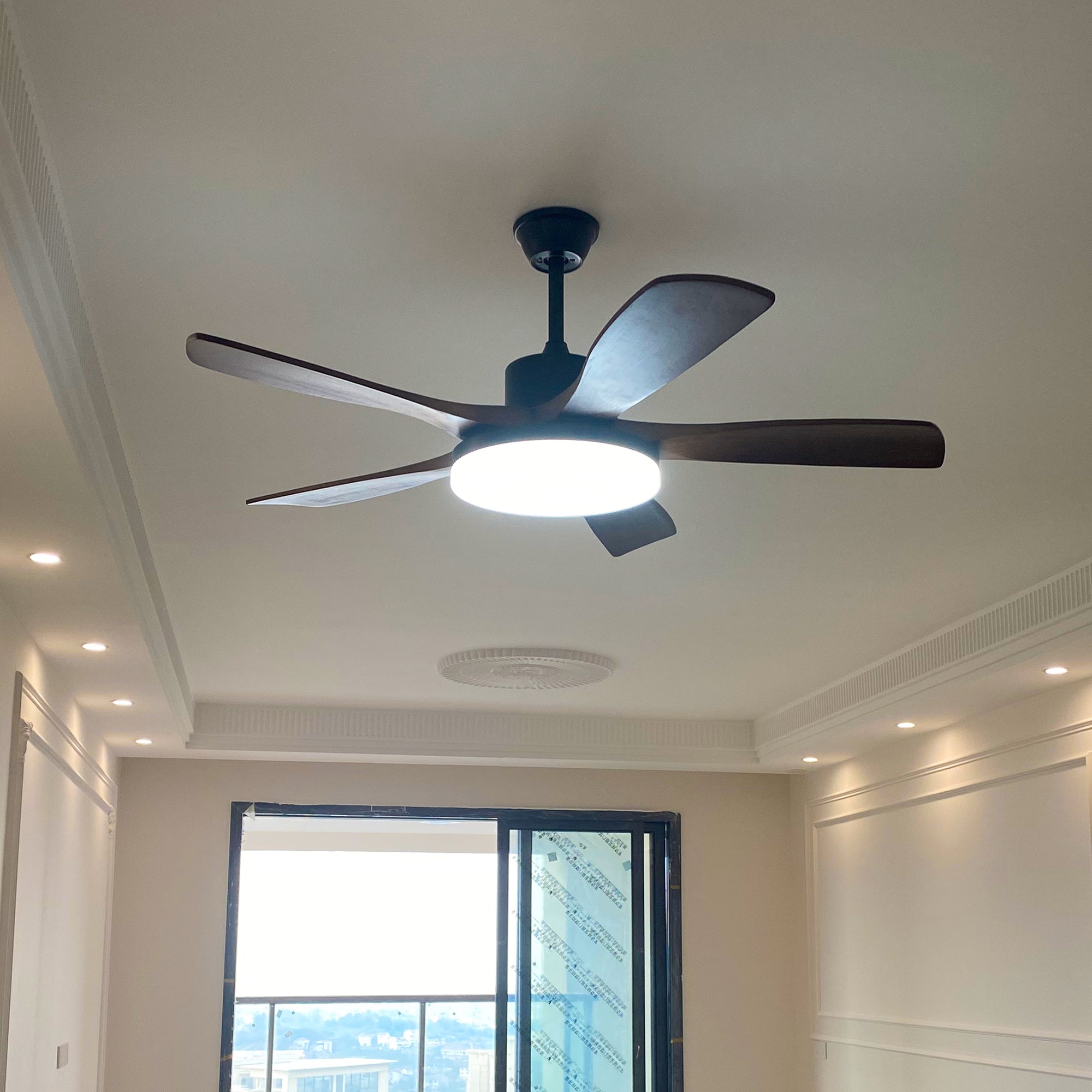 Dark brown five-blade fan with black canopy and housing and a glowing round light in a pale room with a balcony door