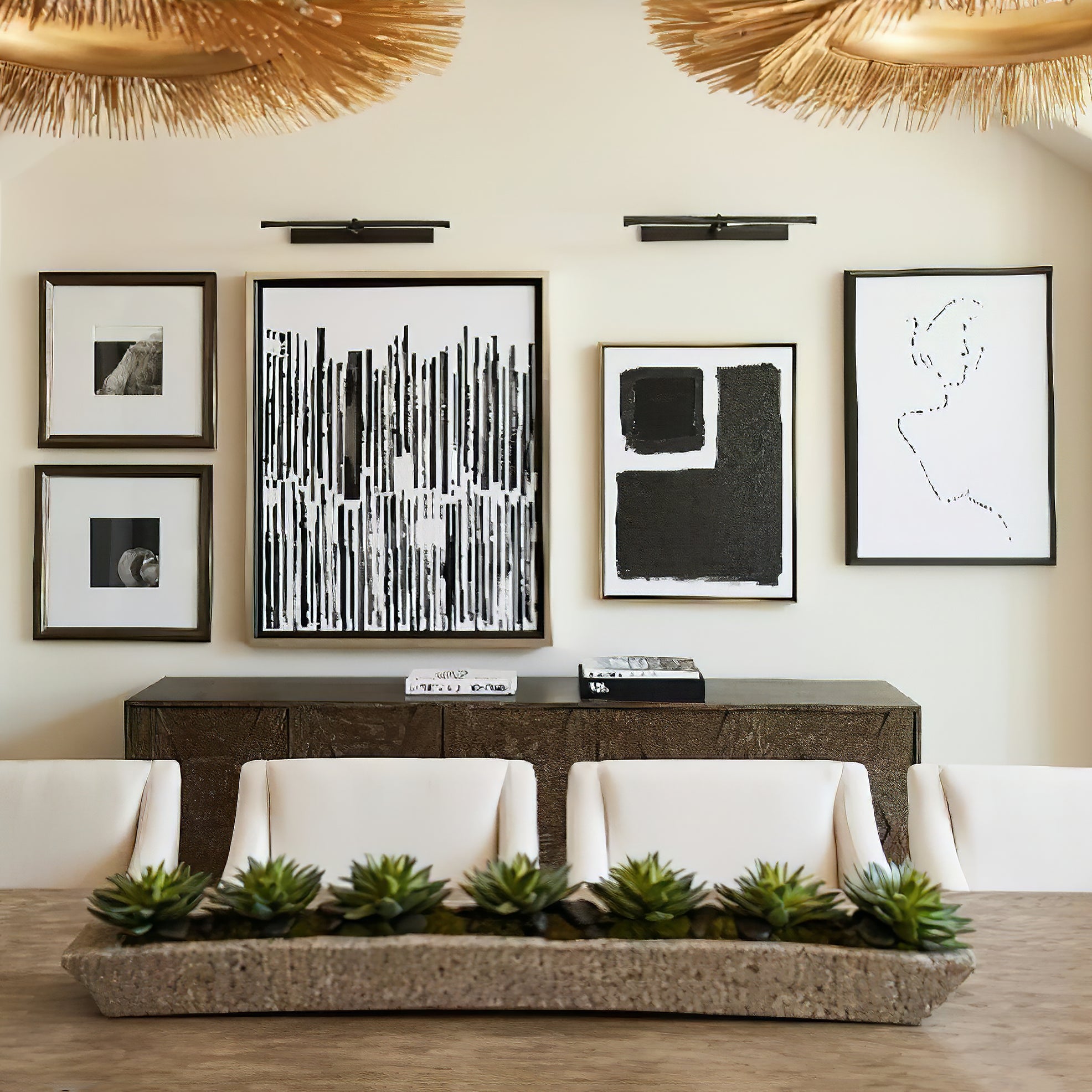 Two black picture lights above framed art beside a dark console, cream chairs and a plant