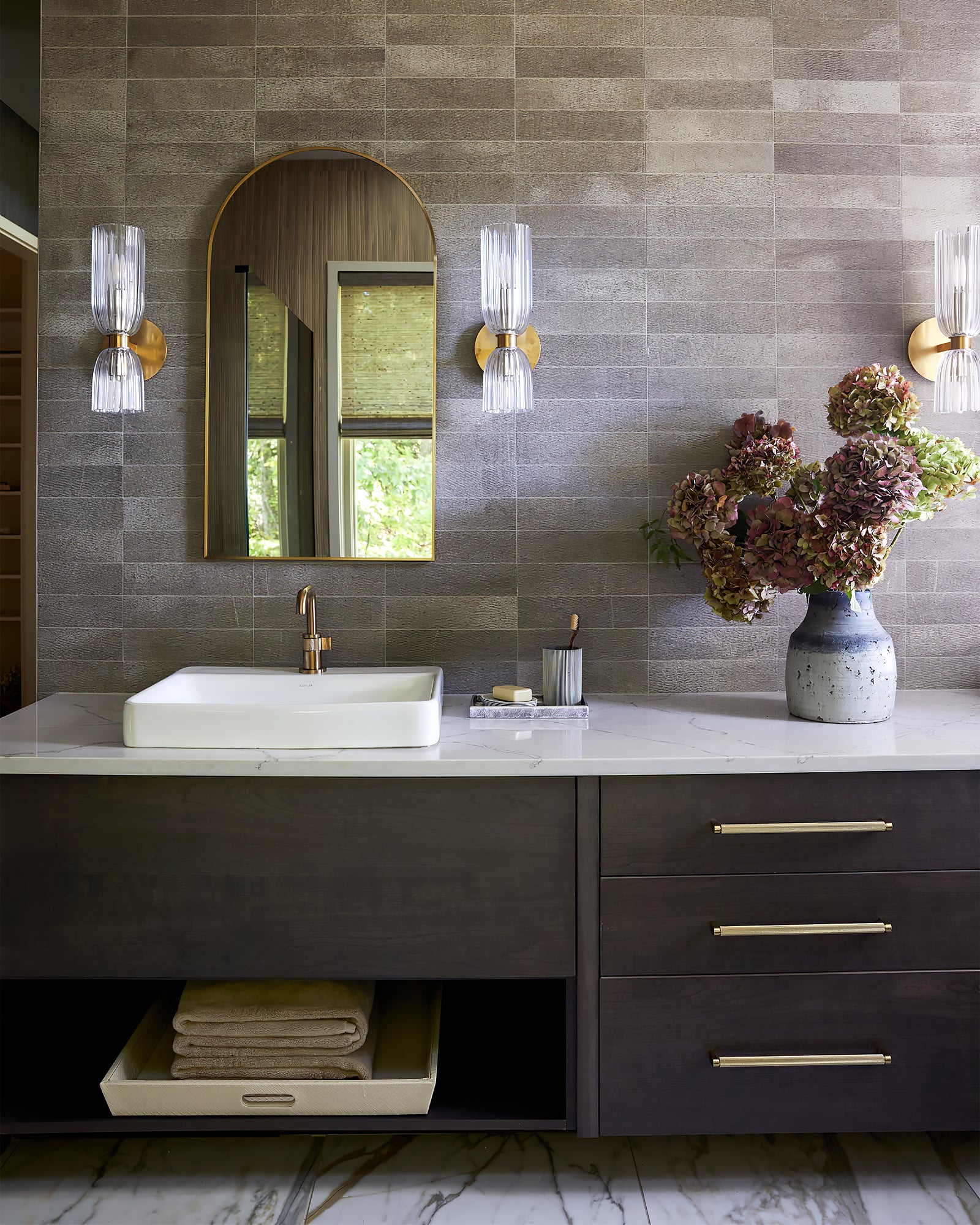 Three double glass sconces visible around an arched mirror and dark-tiled bathroom counter with hydrangeas