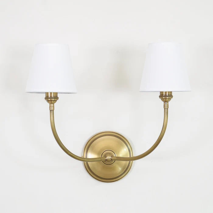 Unlit brass double wall light with tapered white shades against a pale wall.