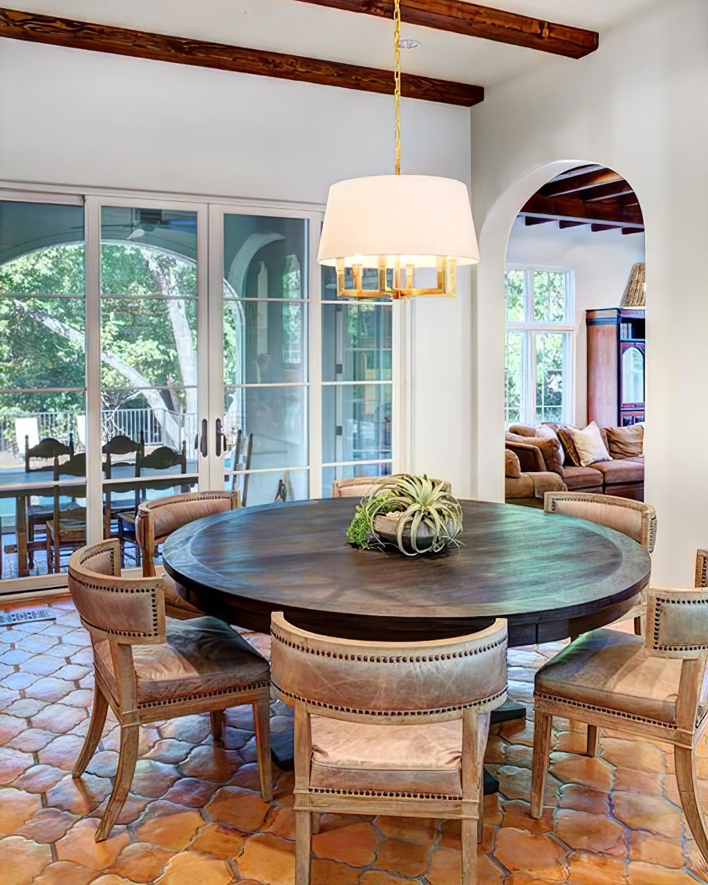 Gold-toned shaded chandelier above a round wooden dining table with upholstered chairs and timber ceiling beams
