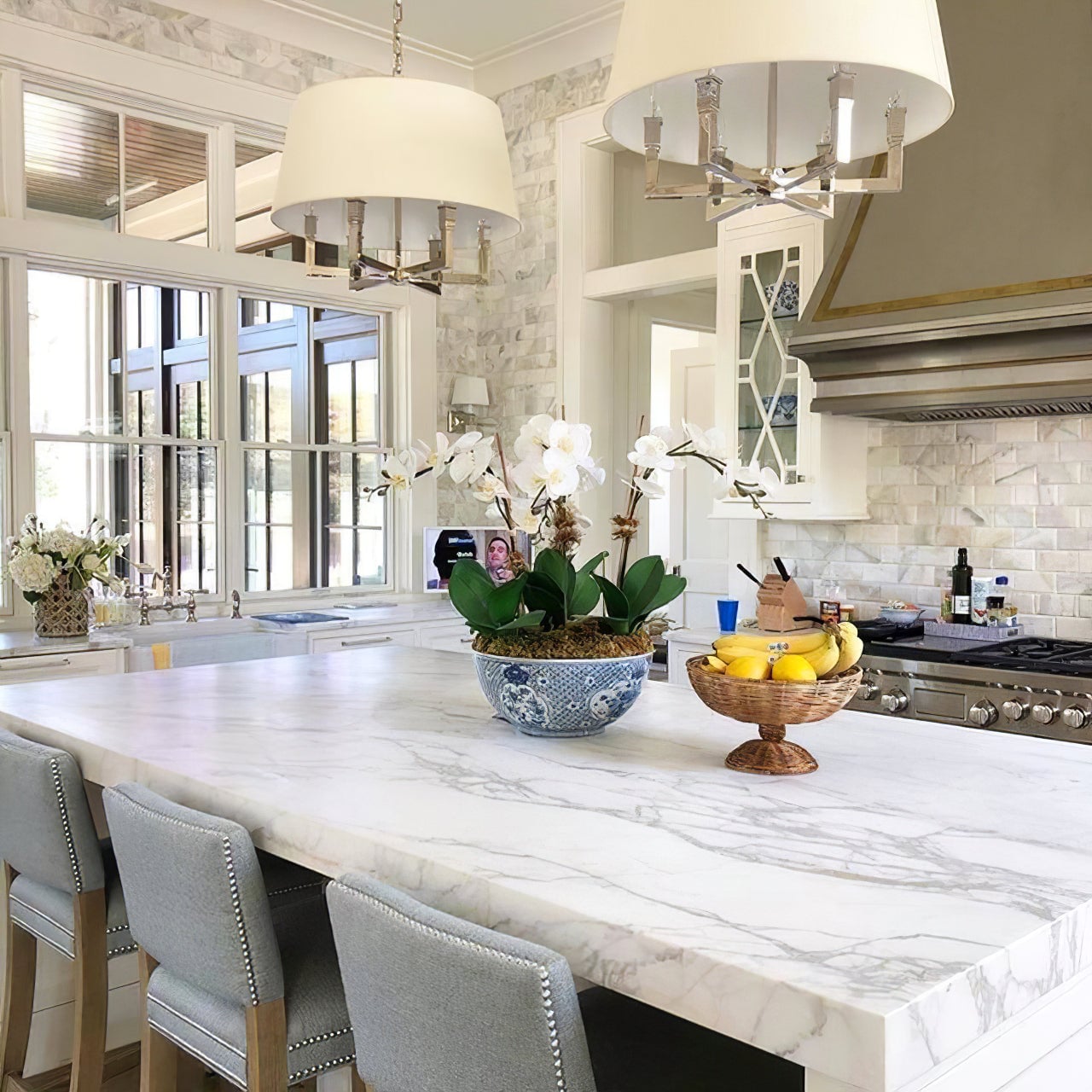 Two silver-looking shaded chandeliers with candle-shaped lamps above an island with orchids and a fruit bowl