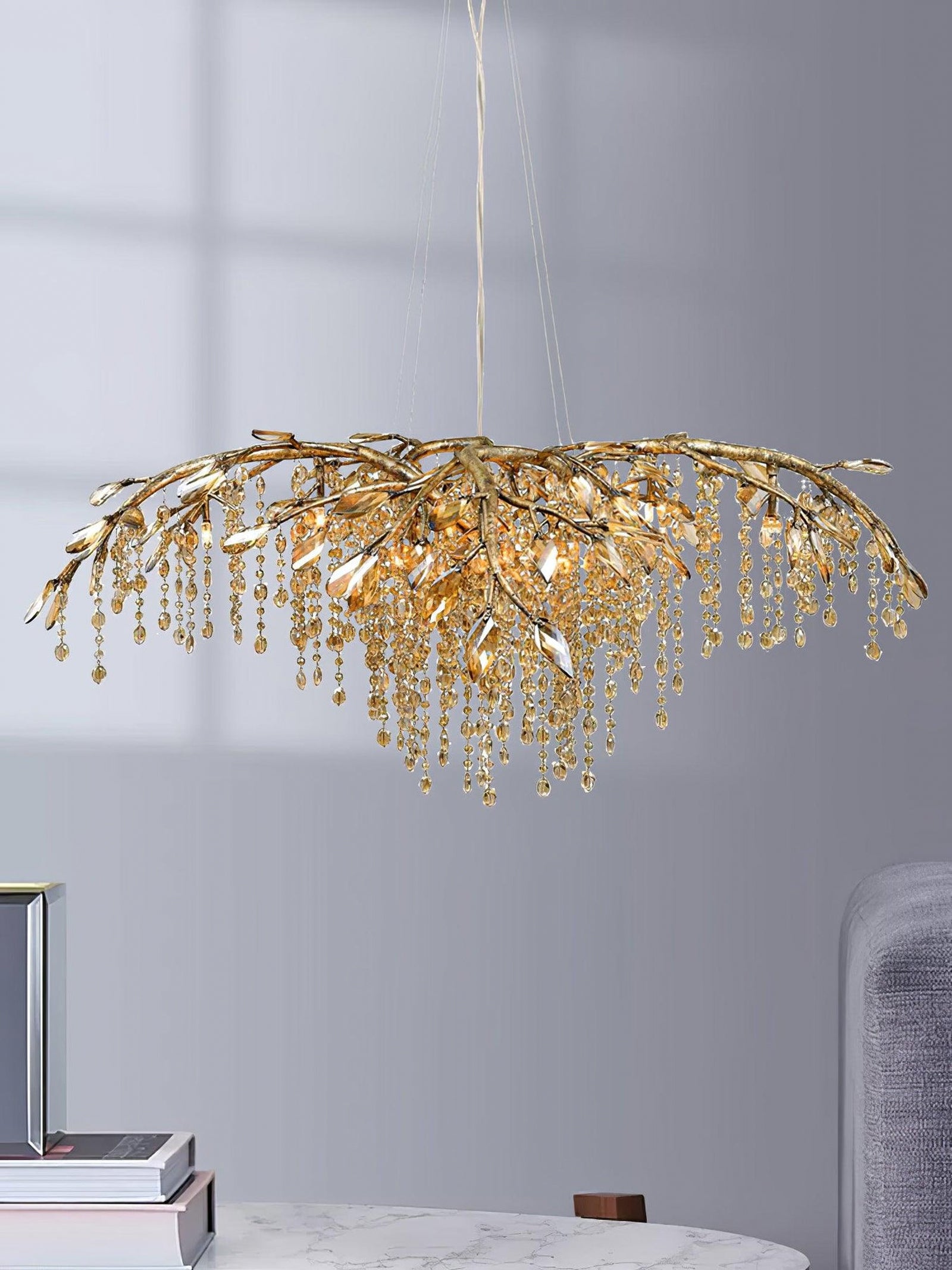 Gold-tone bead chandelier against a gray wall above a partial veined table, books and upholstered seat