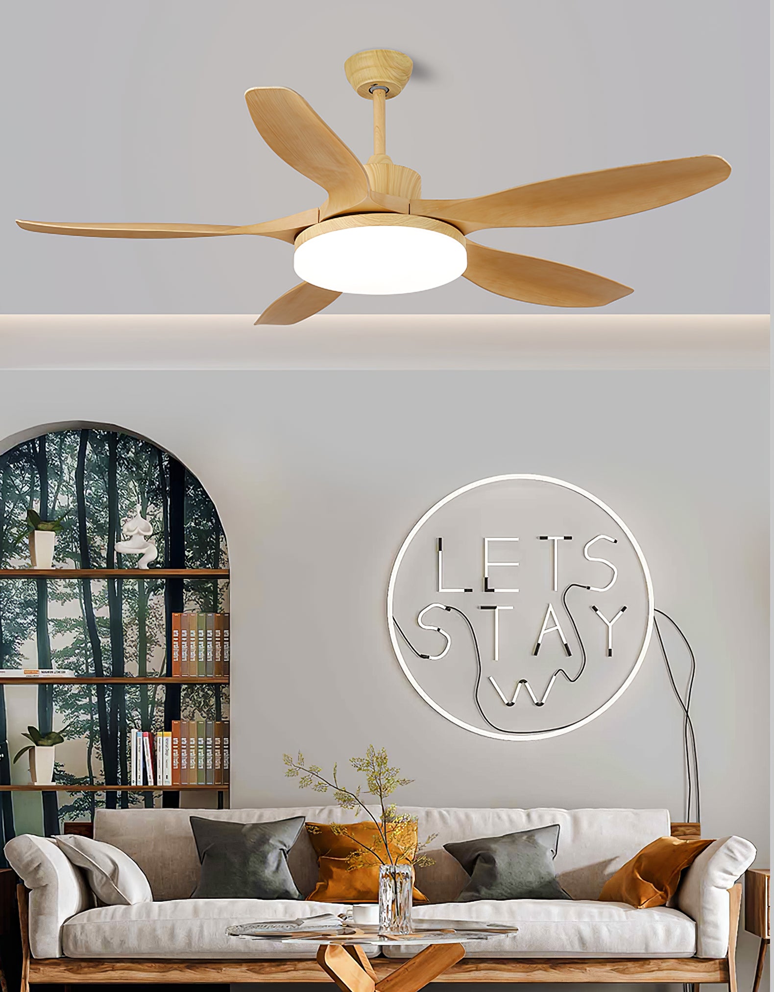 Wood-tone five-blade fan with a round light above a cream sofa with grey and ochre cushions beside an arched bookcase