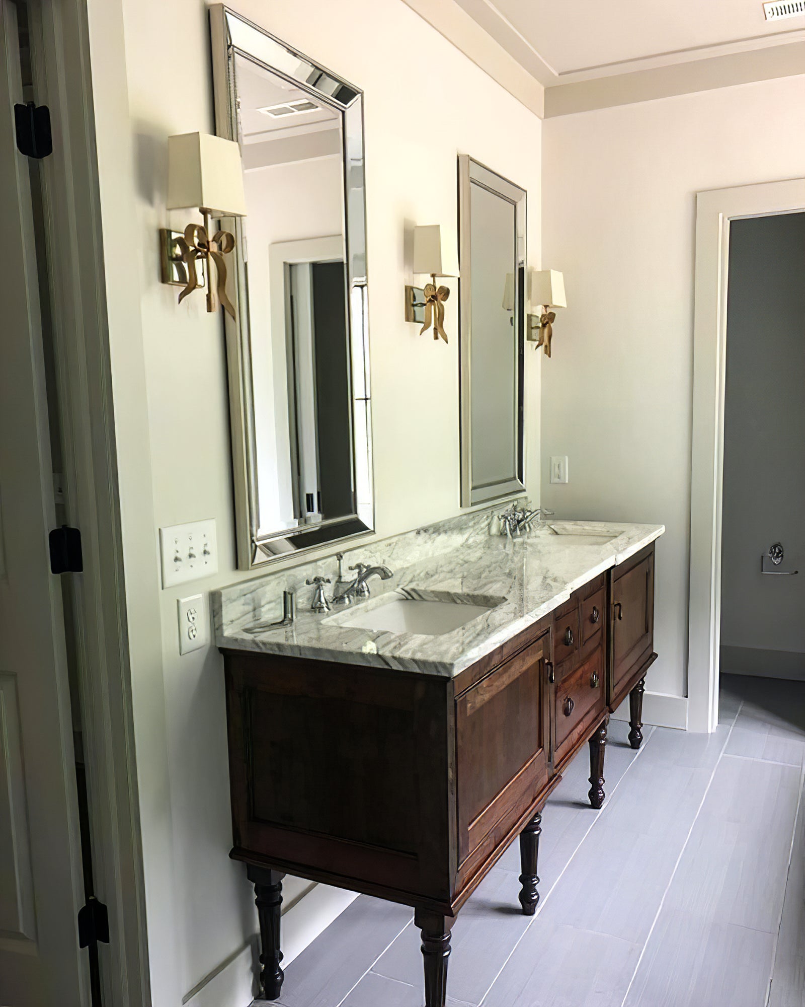 Three brass-tone bow lights with pale shades arranged around two mirrors above a dark wood double-basin cabinet