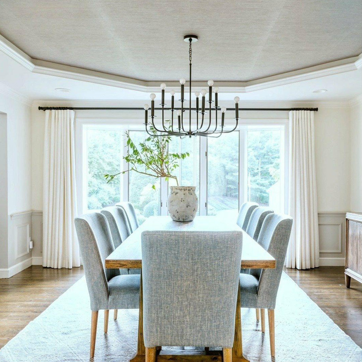 Black multi-arm chandelier above a rectangular dining table with gray chairs, a plant vase and large window